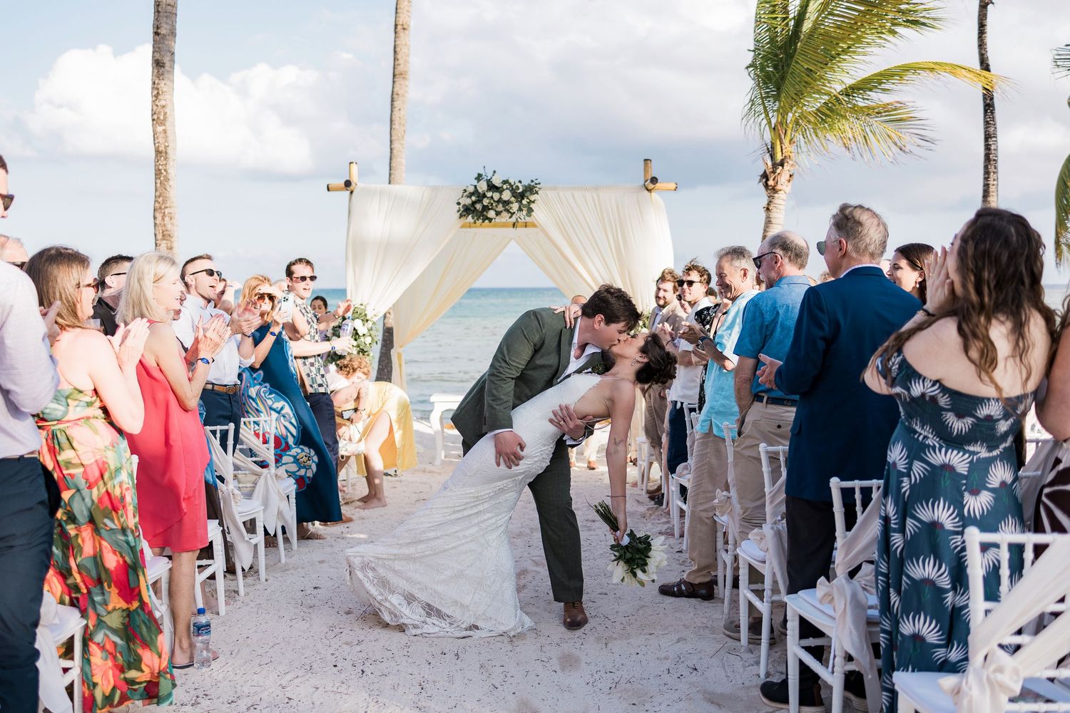 Couple shares romantic dip kiss at outdoor beach wedding ceremony with guests cheering, palm trees, and white draped arch.
