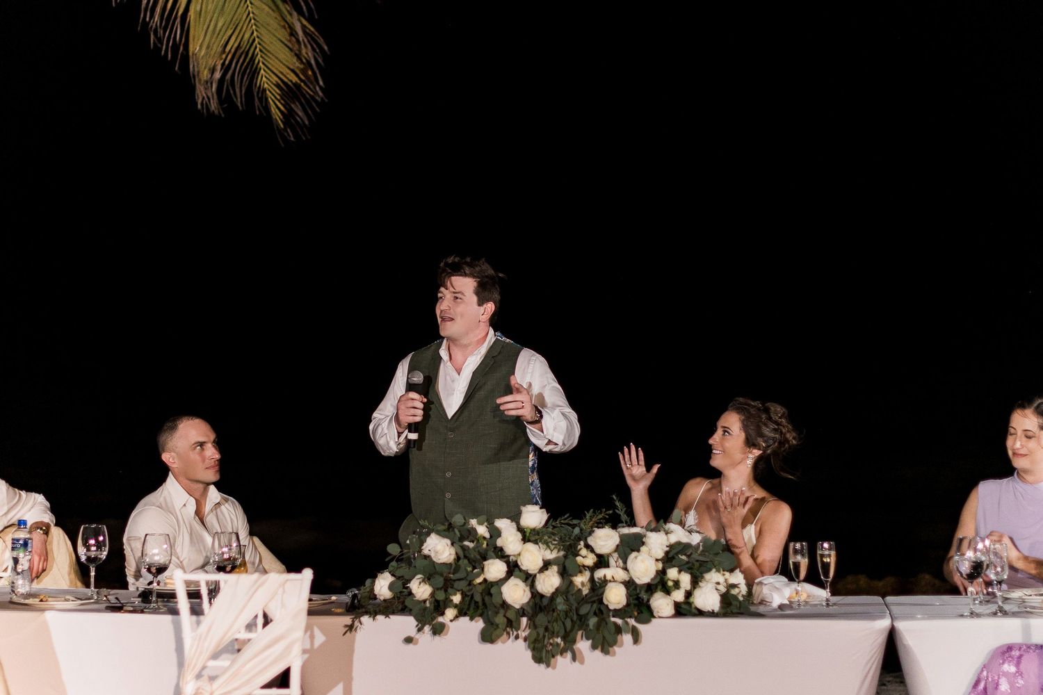 A groom gives a heartfelt wedding speech at an outdoor reception table adorned with white roses under a night sky.