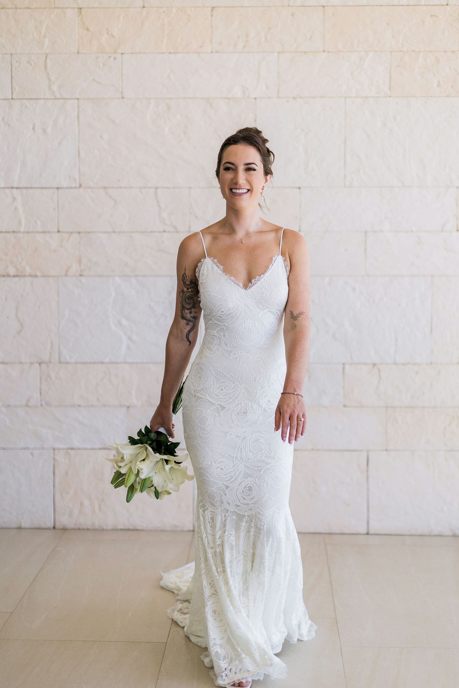 Smiling bride in white lace spaghetti strap wedding gown holding white bouquet, standing against stone wall.