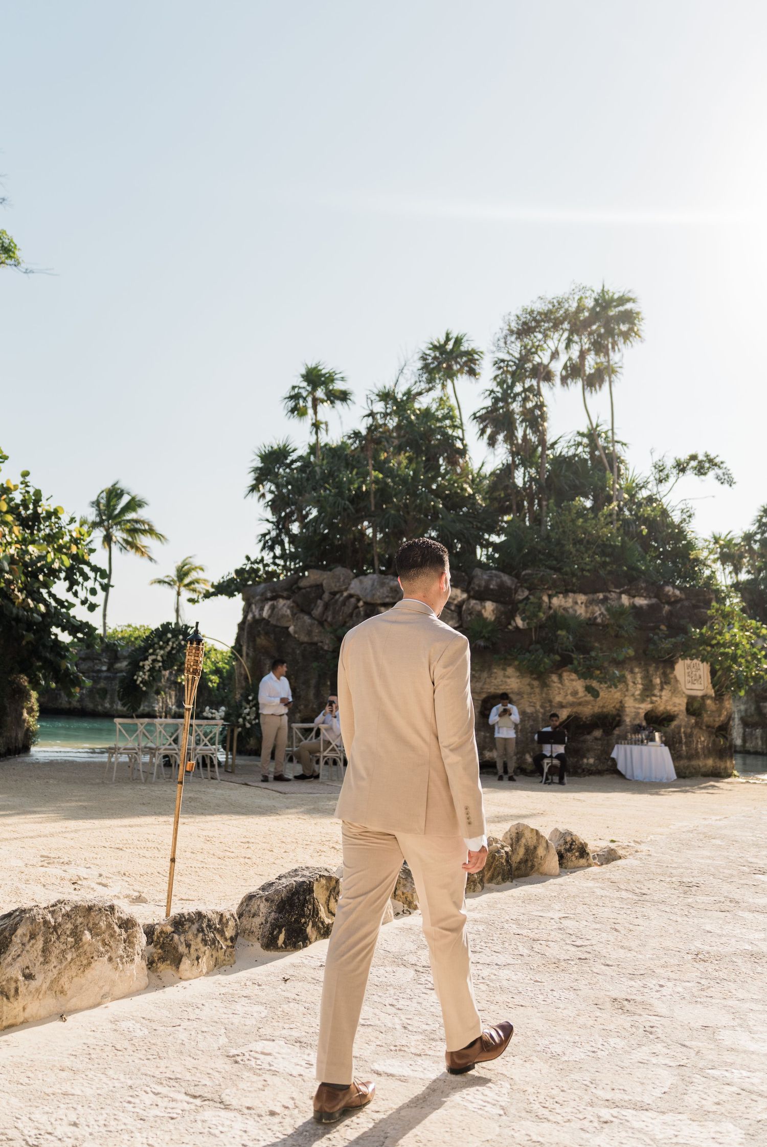Xcaret Arte Wedding - Groom in beige suit walking toward tropical beach wedding ceremony with palm trees and rocky backdrop.