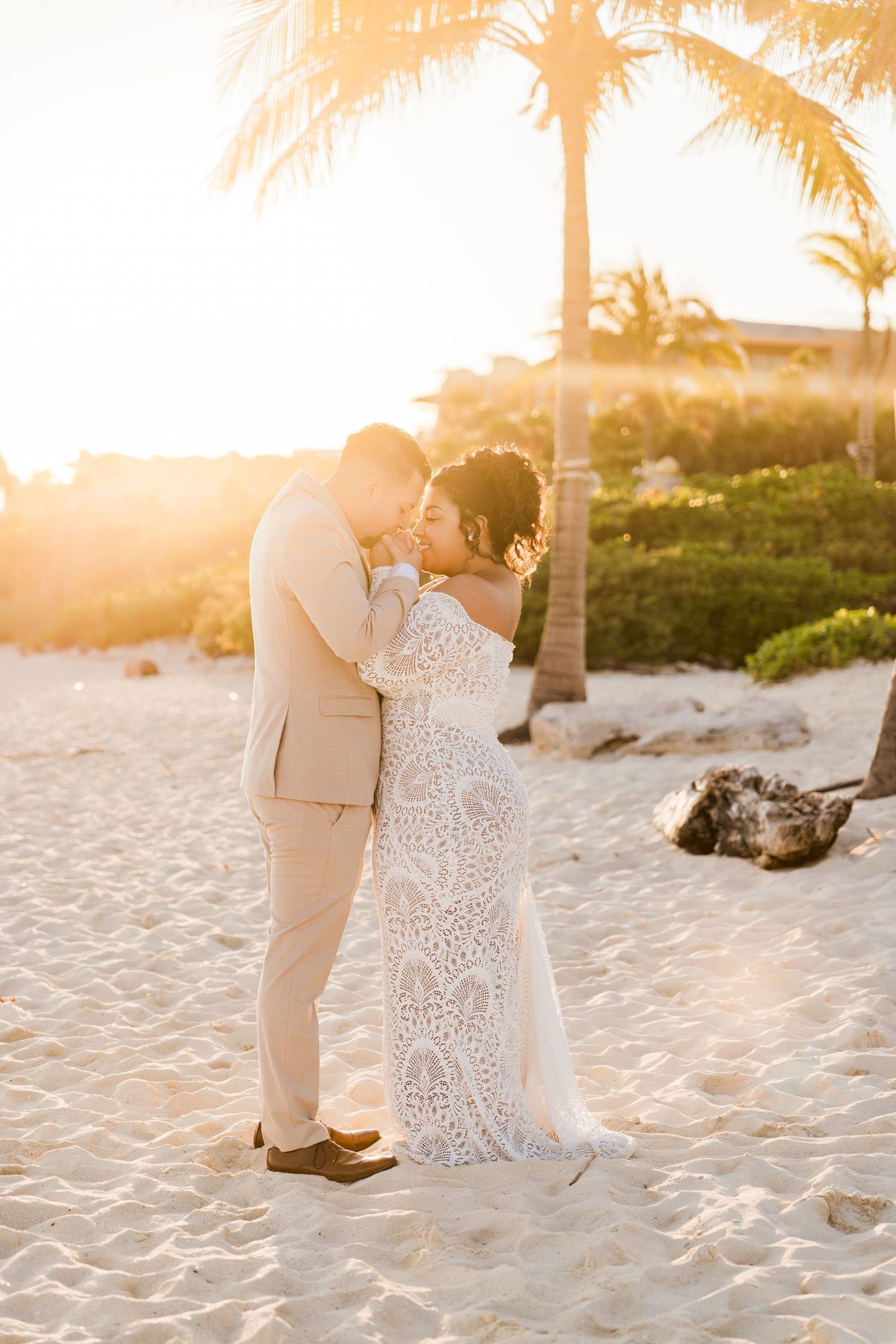 Xcaret Arte Wedding - Bride and groom share romantic sunset kiss on white sand beach, bride in lace gown, groom in tan suit