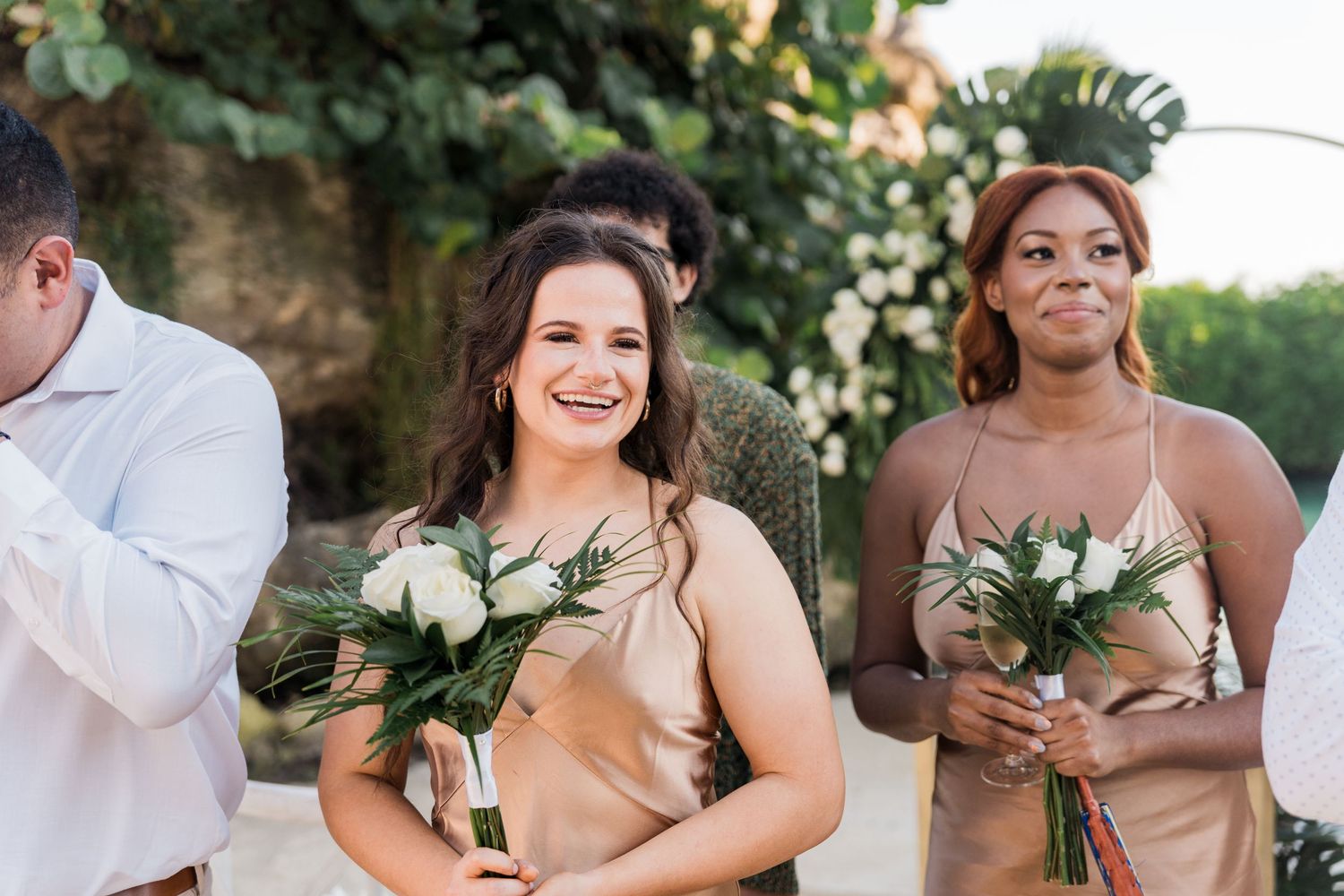 Xcaret Arte Wedding - Two smiling bridesmaids in gold dresses holding white bouquets stand at an outdoor wedding ceremony.