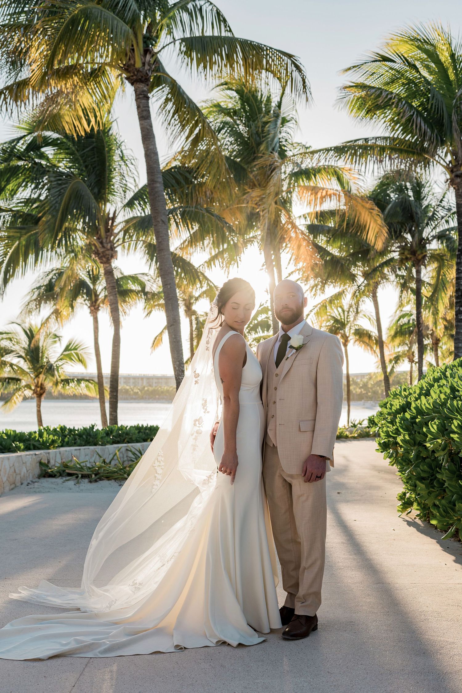 Bride and groom pose among tropical palm trees at sunset in a beachside wedding portrait.