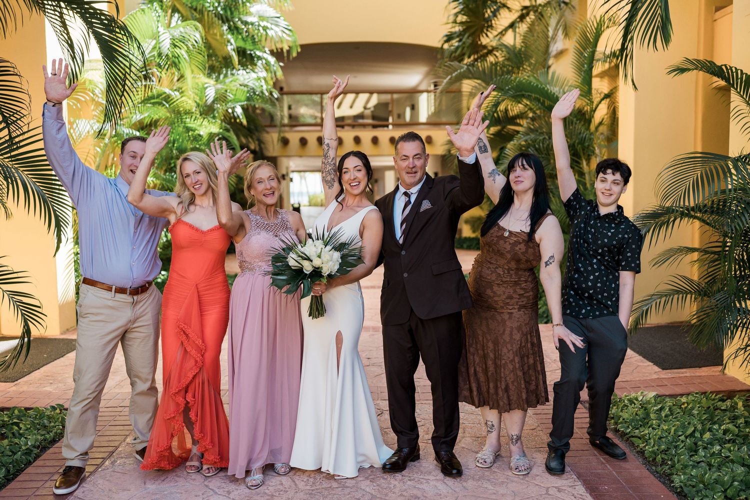 Wedding party celebrating outdoors, bride in white gown holding bouquet, guests in colorful formal attire, arms raised joyfully.