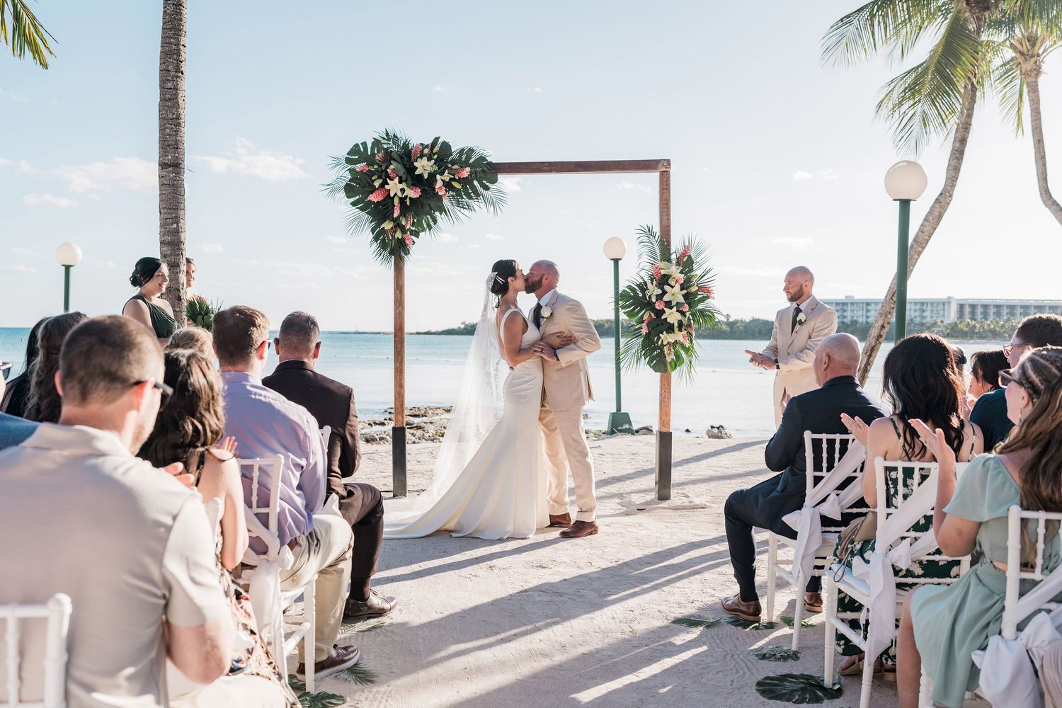 Couple kissing at outdoor beach wedding ceremony with floral arch, white chairs, and guests seated by the ocean.