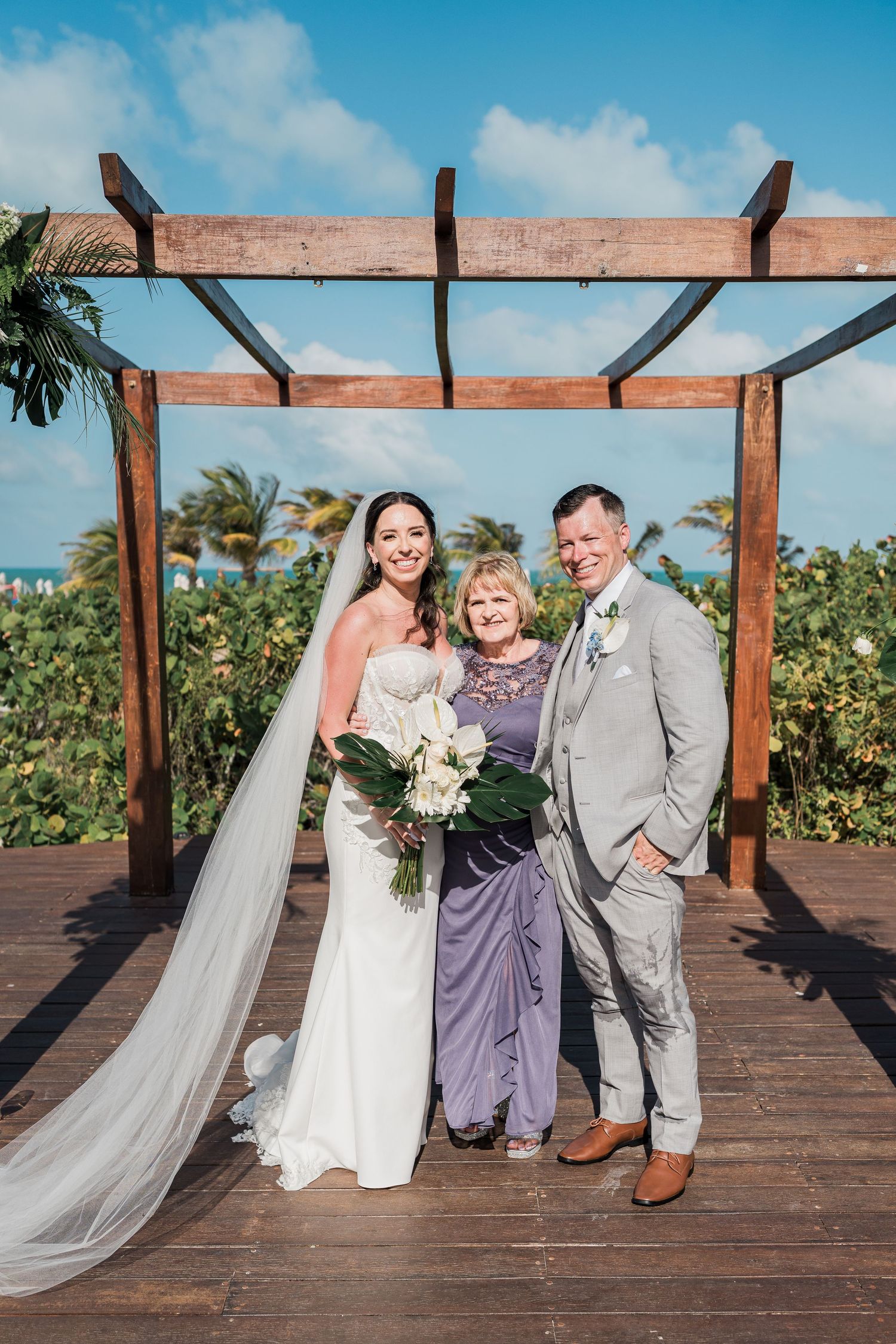 Bride in white gown and groom in gray suit pose with older woman in purple dress under wooden pergola by the ocean.