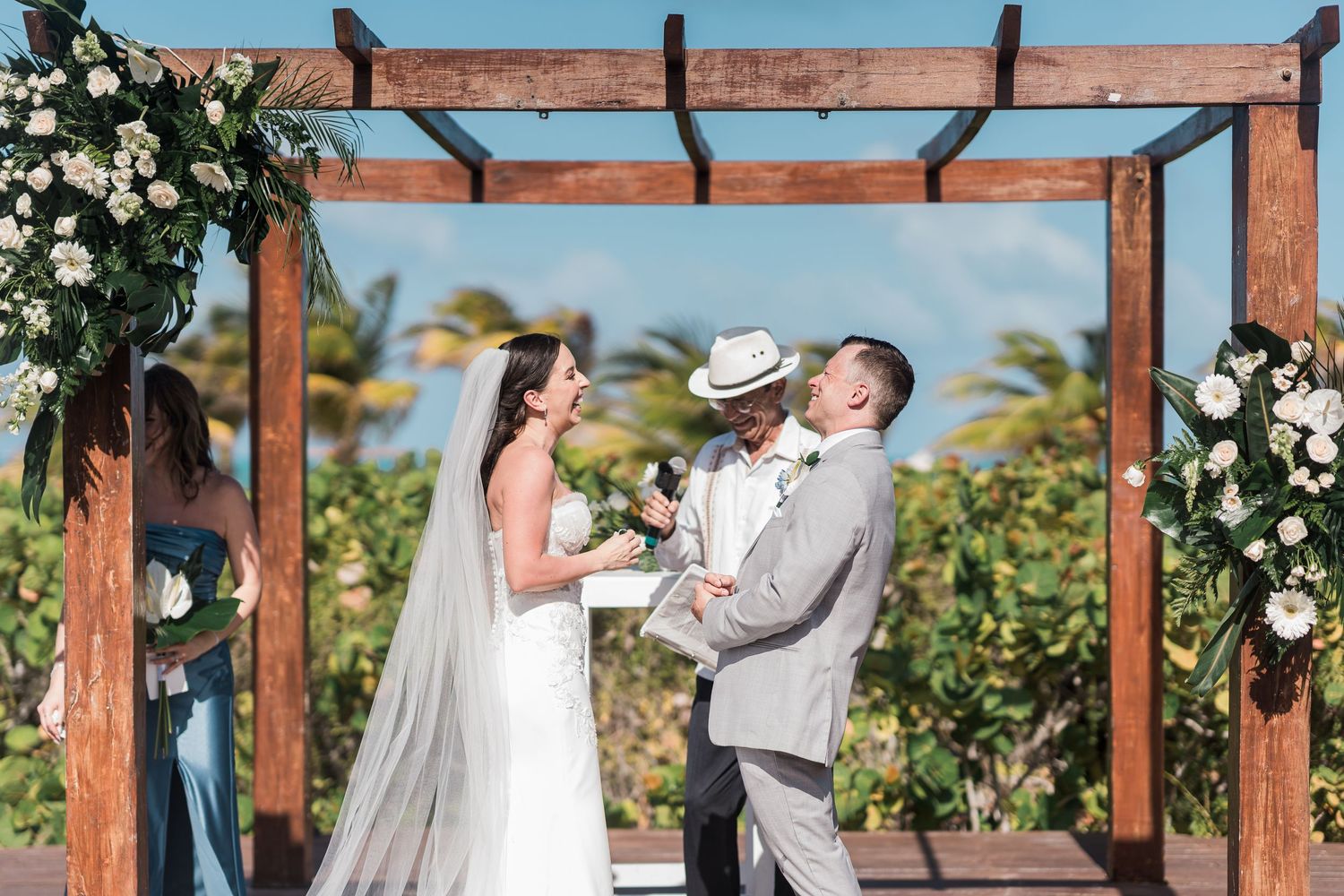 Couple exchanging vows under wooden arch with white floral decor at outdoor beach wedding ceremony.