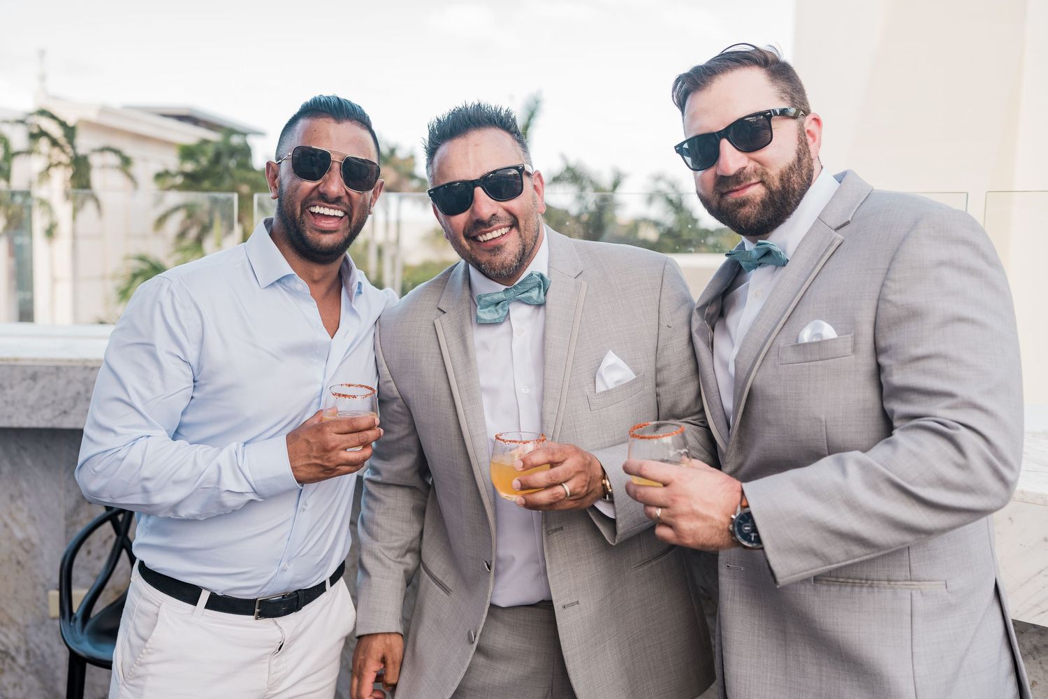 Three men in gray suits and bow ties smiling and holding drinks outdoors at a formal event.