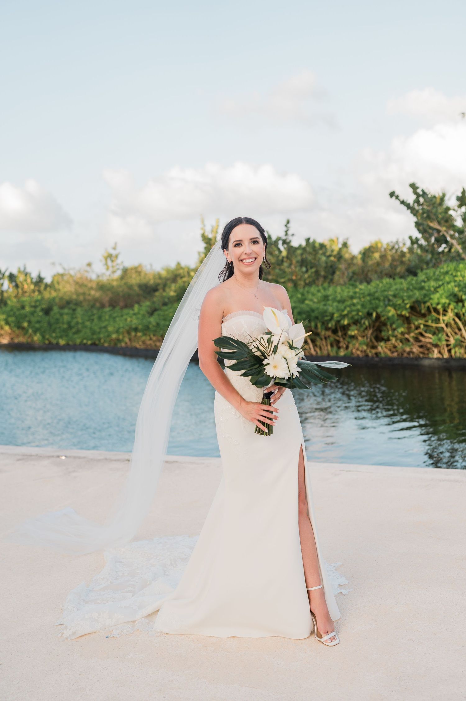 Bride in strapless white gown with veil holding bouquet by tropical waterway on sunny day.