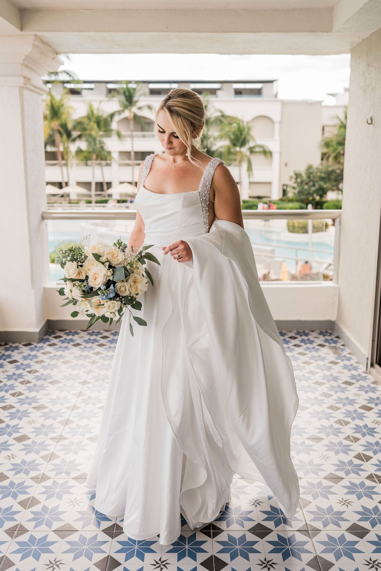 Bride in white gown holding bouquet on tiled balcony with palm trees and pool in tropical resort. Hard Rock Riviera Maya wedding