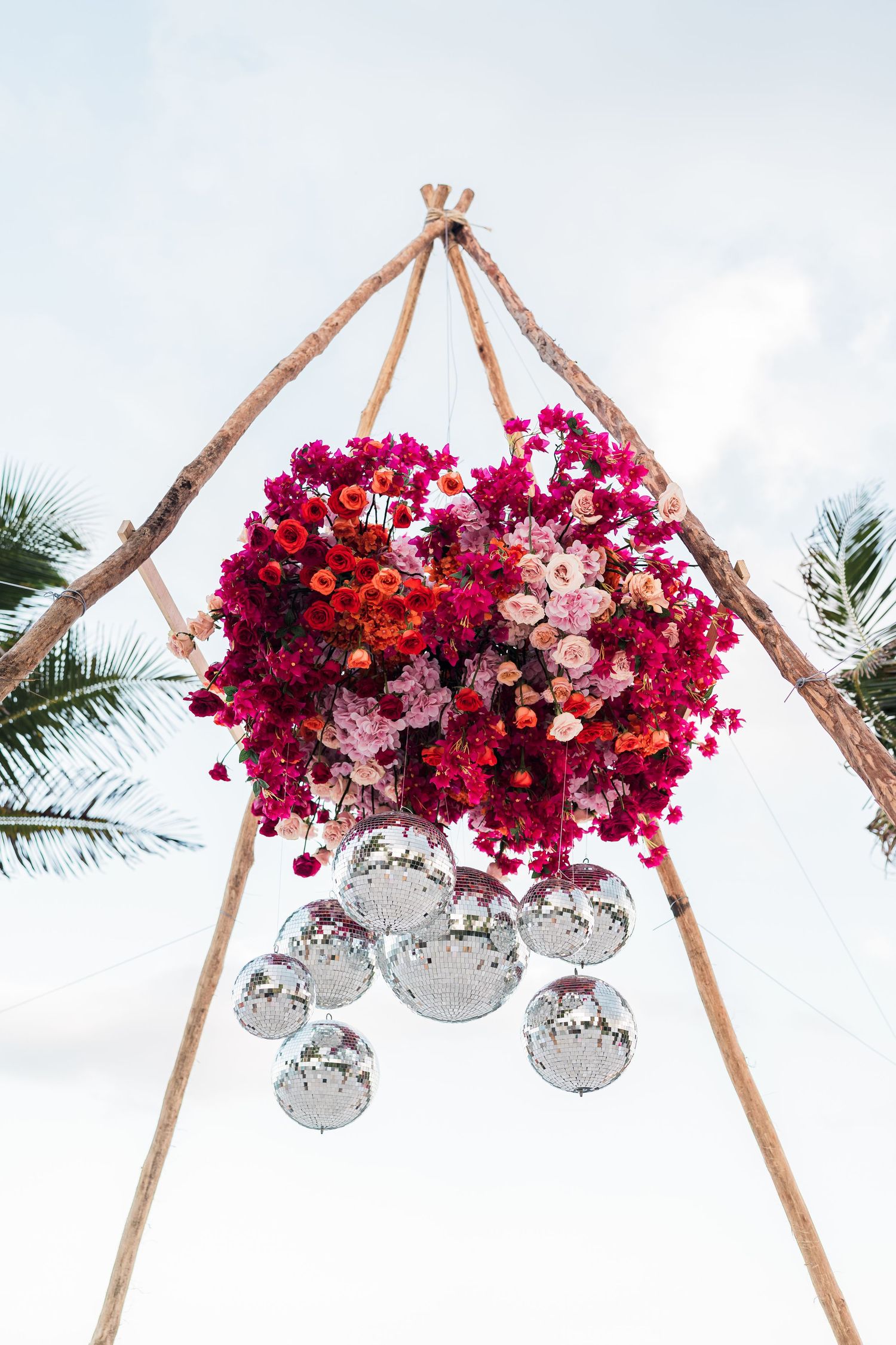 Floral chandelier with red and pink blooms and hanging disco balls suspended from rustic wooden frame outdoors.