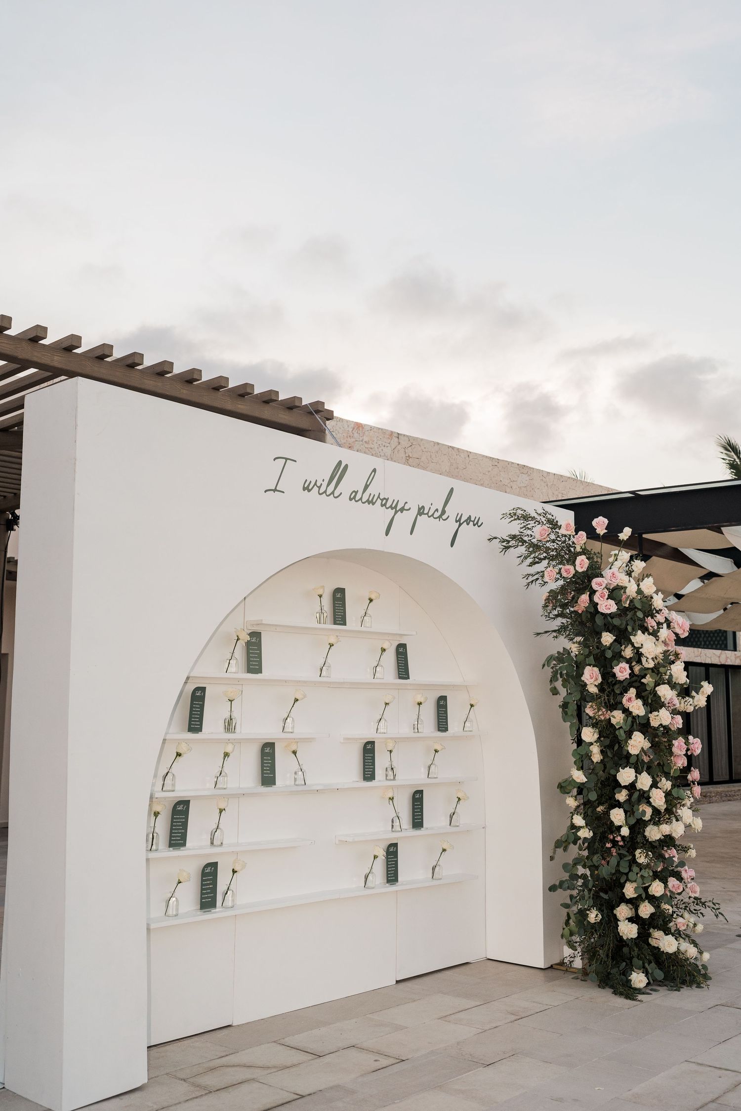 White arched wedding escort card display with floral arrangement and cursive quote at outdoor reception.