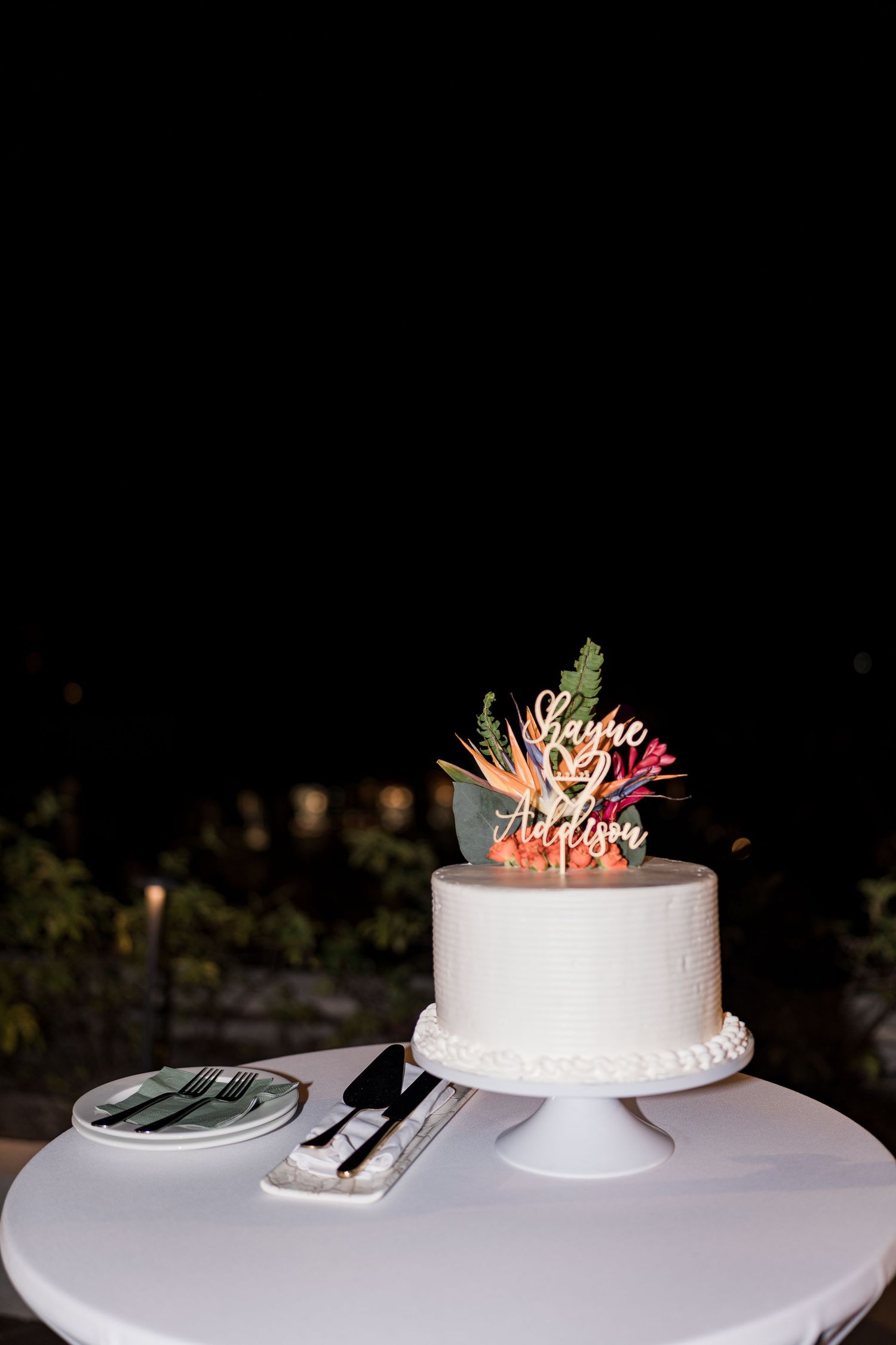Elegant single-tier white wedding cake with tropical floral topper and custom name sign on a white pedestal stand.