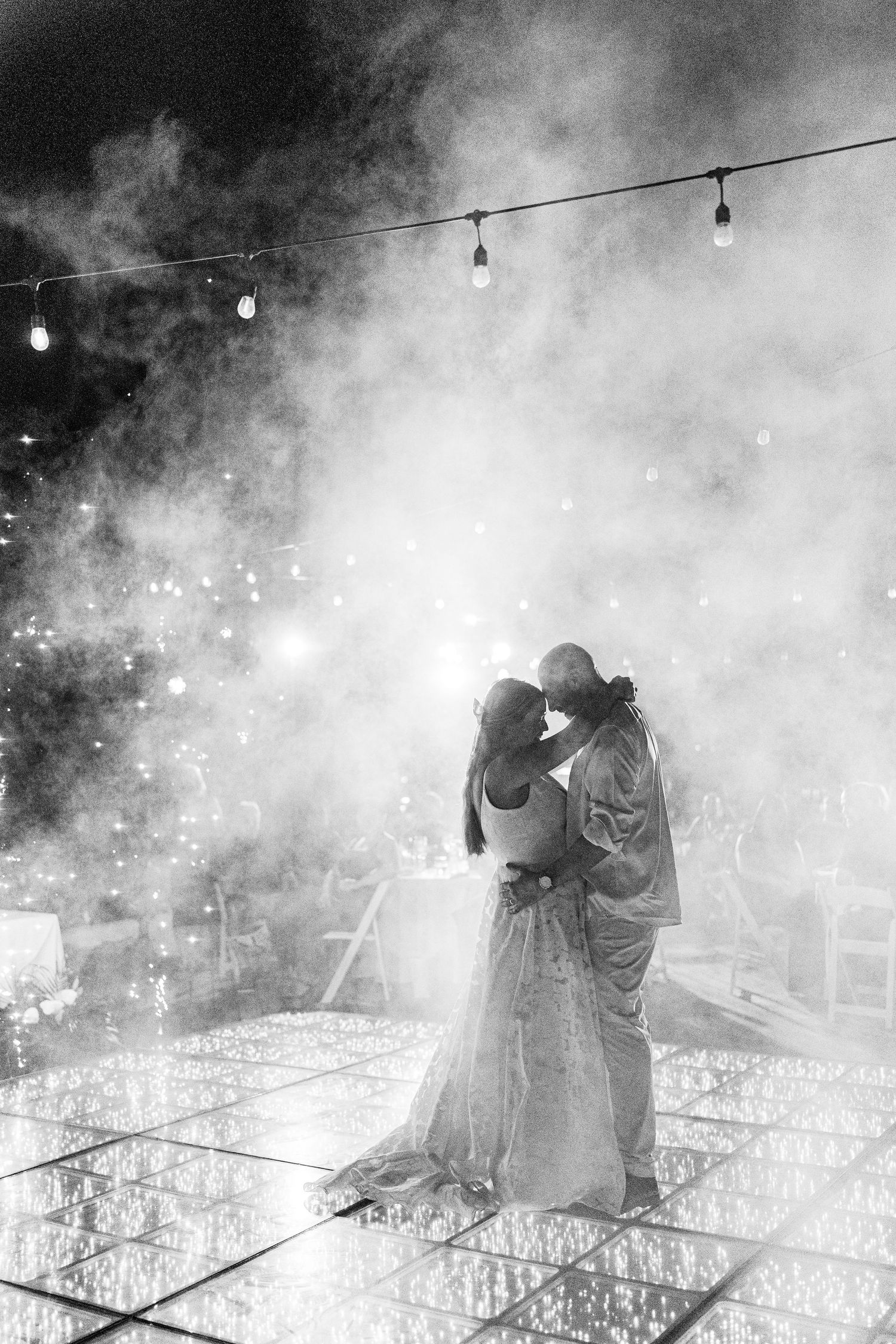 Black and white photo of bride and groom sharing first dance on illuminated floor surrounded by dramatic fog at wedding.