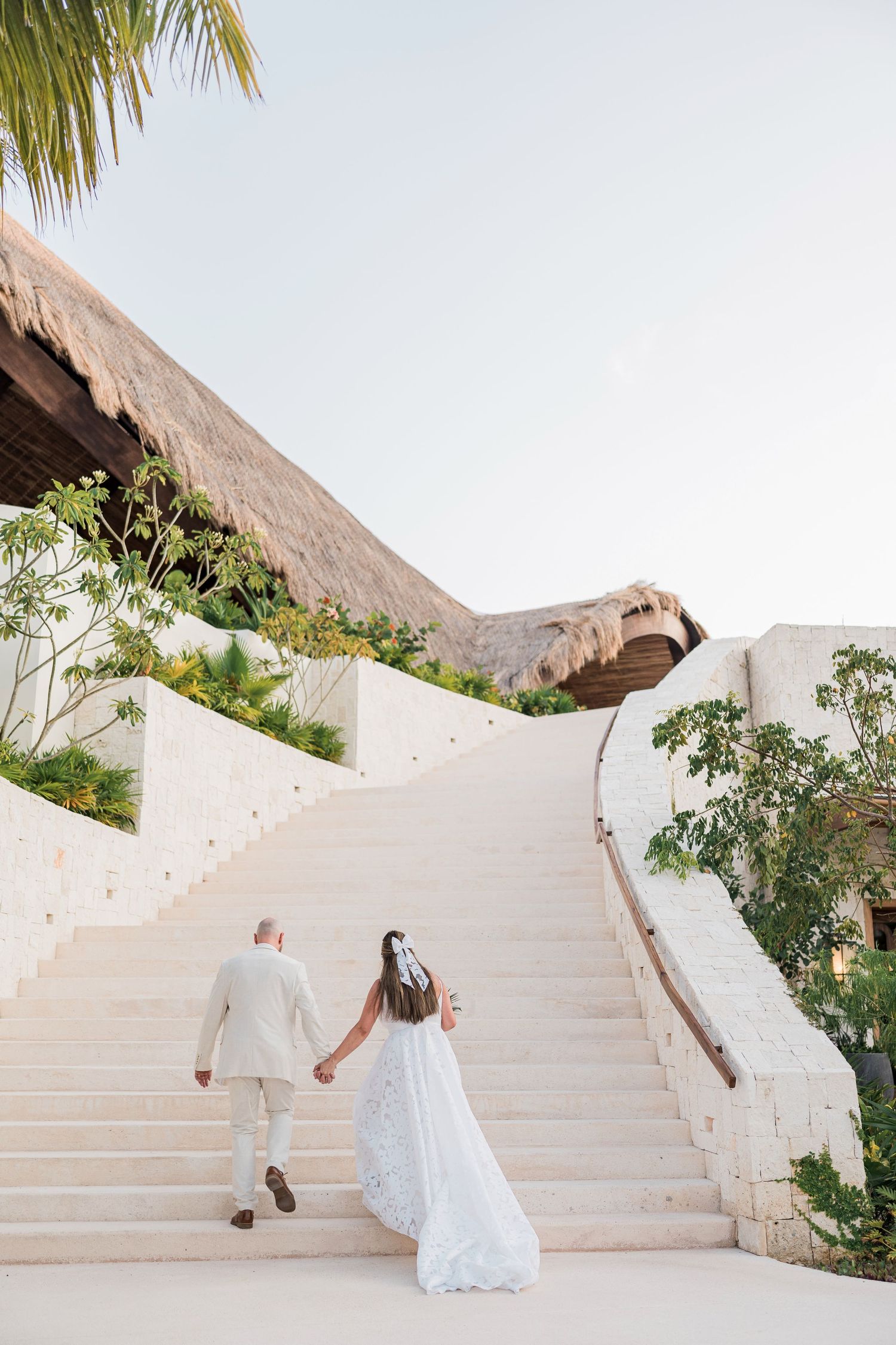 Bride and groom holding hands walking up white stone stairs at a tropical resort wedding venue.