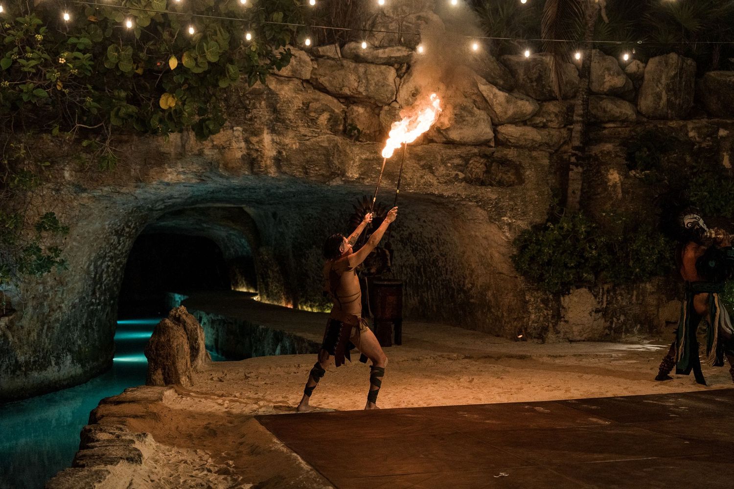 Fire performer balances flaming torch at night in a cave-like venue with string lights and sandy floor.