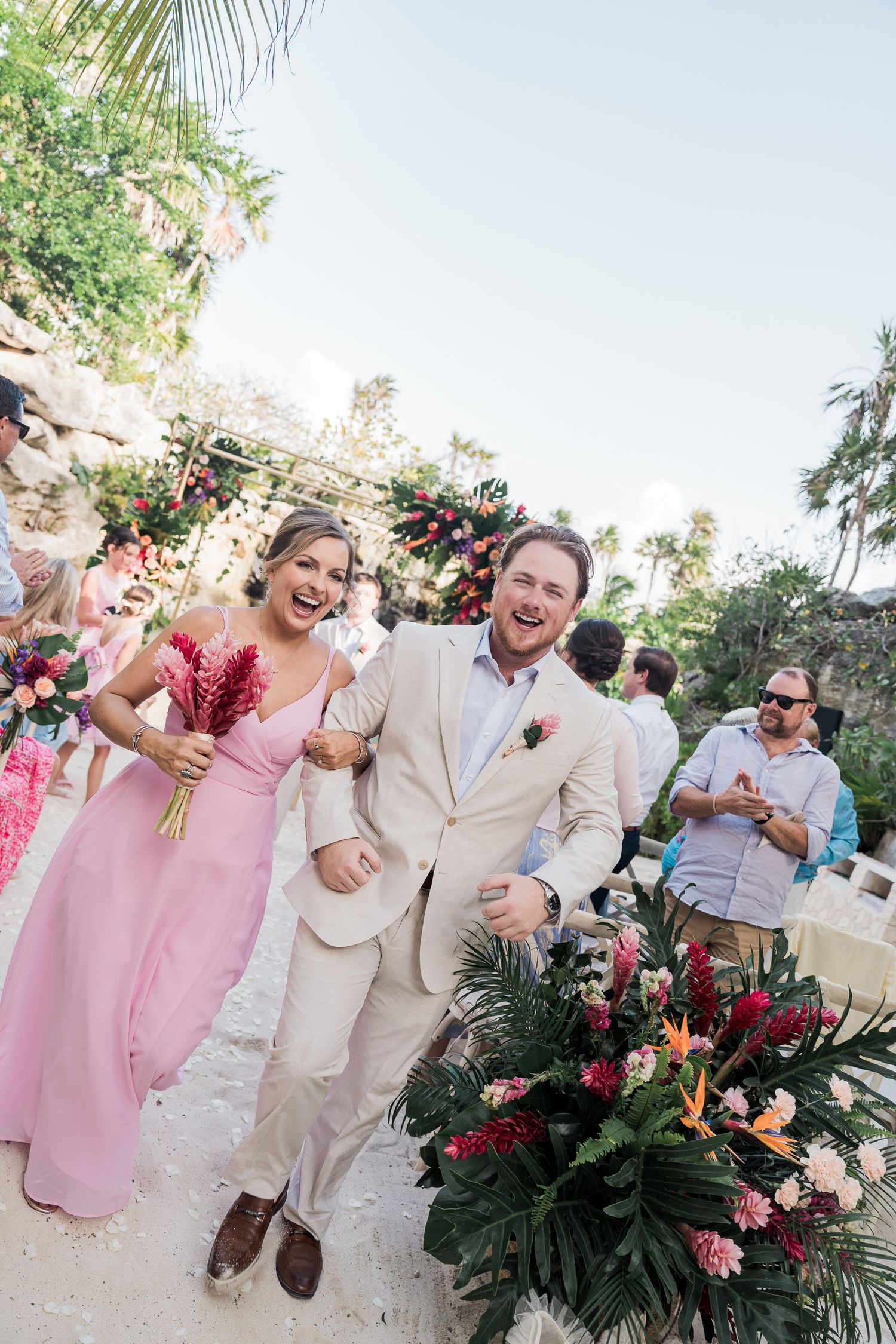 Joyful bride in pink dress and groom in tan suit walk down tropical wedding aisle with colorful floral arrangements.