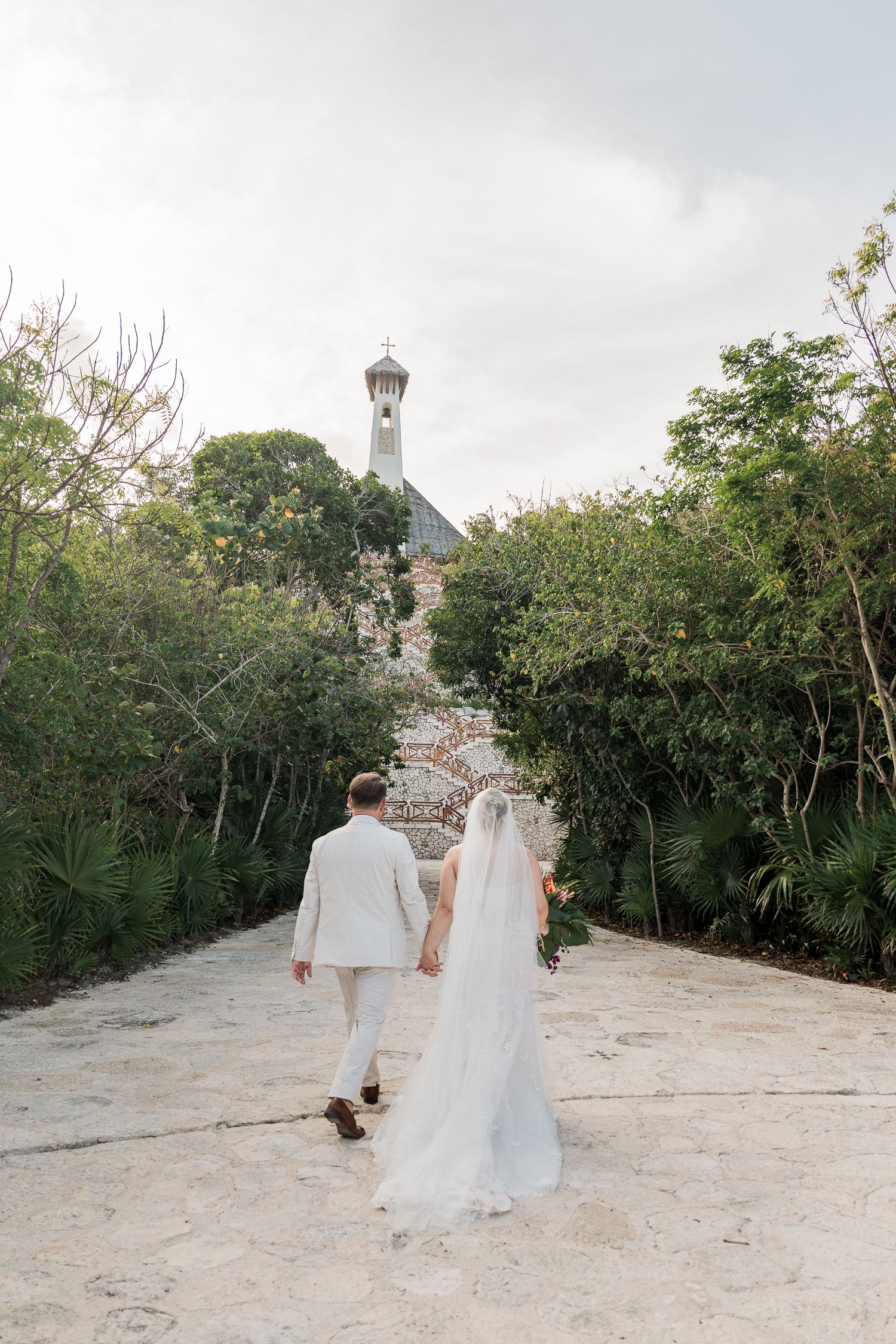 Bride and groom walking hand in hand toward a chapel surrounded by lush greenery on their wedding day.