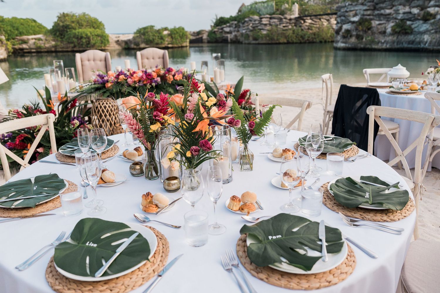 Tropical beach wedding reception table with palm leaf plates, wicker chargers, lush floral centerpiece, and crystal glassware by the water.