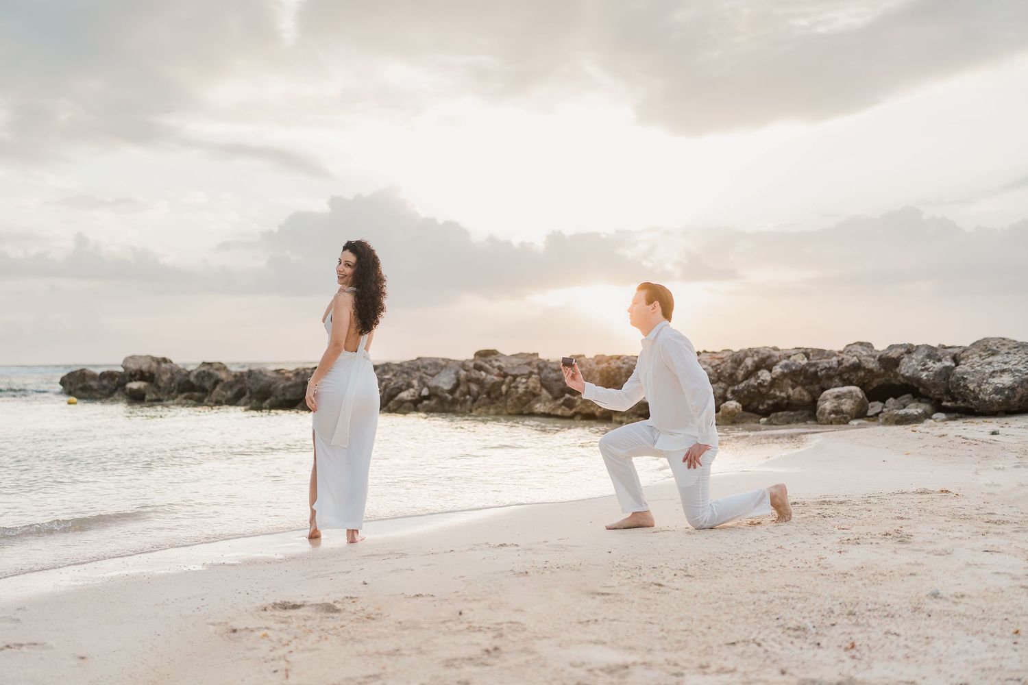 Man proposing on one knee to woman in white dress on tropical beach at sunset.