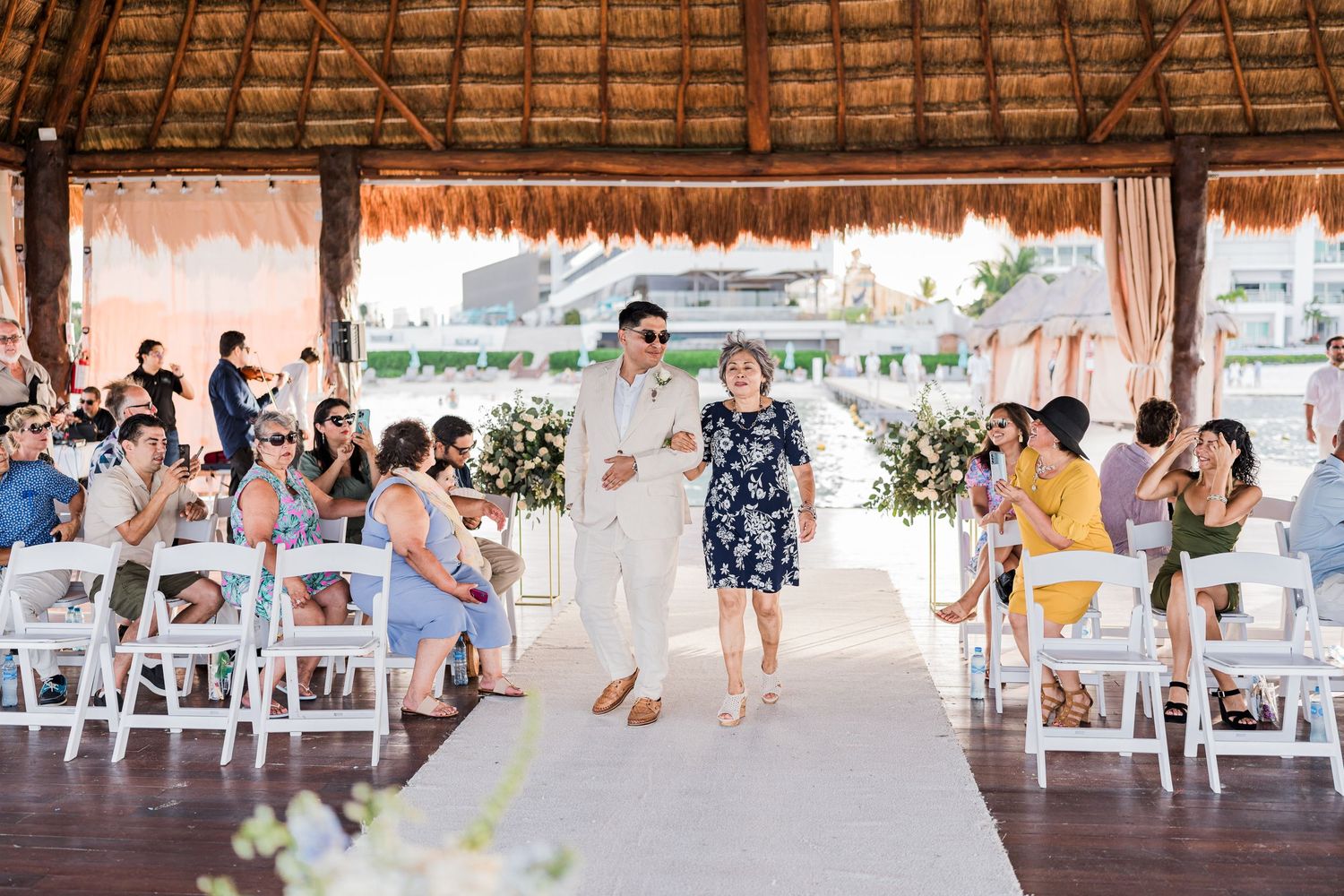 Groom escorted down aisle by mother at outdoor beach wedding ceremony.