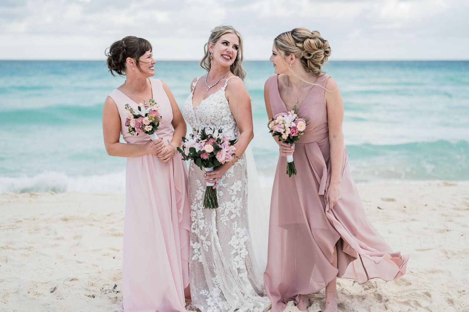 Bride and two bridesmaids in pink dresses holding bouquets on a tropical beach.