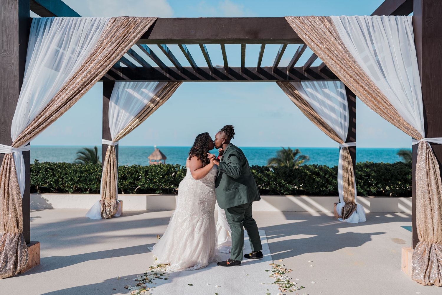 Newlyweds share first kiss under a draped pergola with turquoise ocean backdrop.