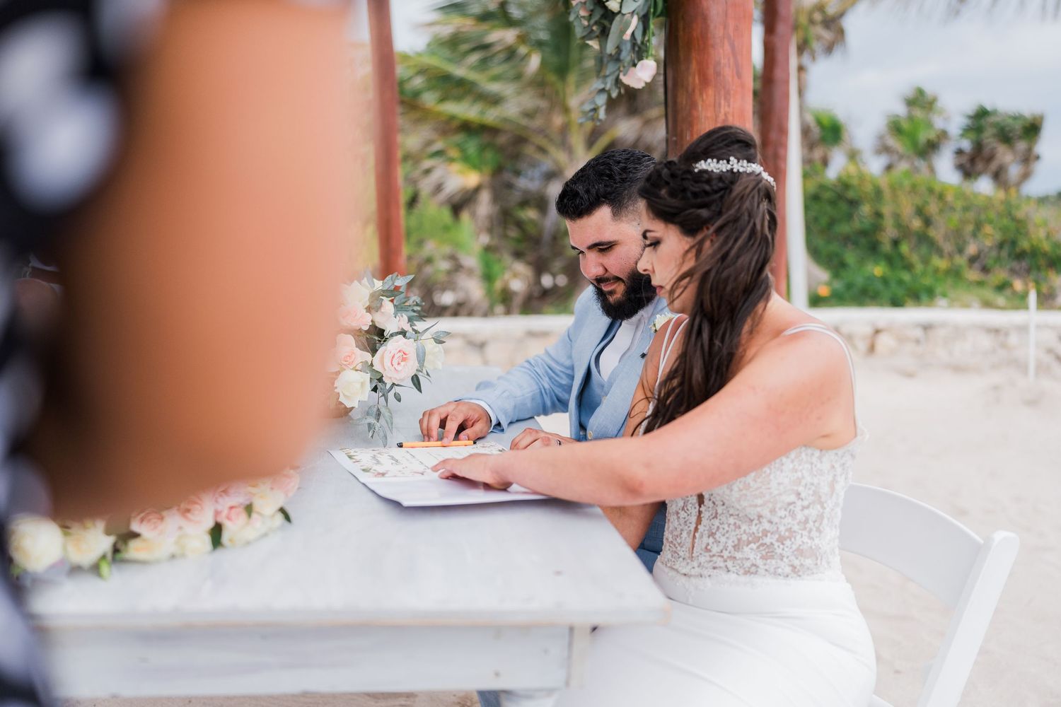 Newlyweds sign marriage certificate together at white table during beach wedding ceremony.