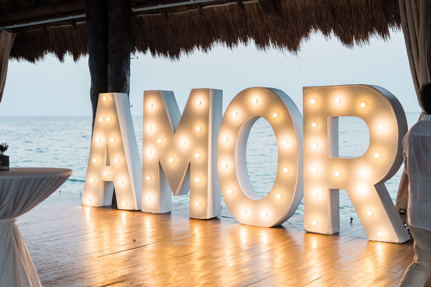 Illuminated AMOR marquee letter sign glowing at a romantic beach wedding reception under a palapa.