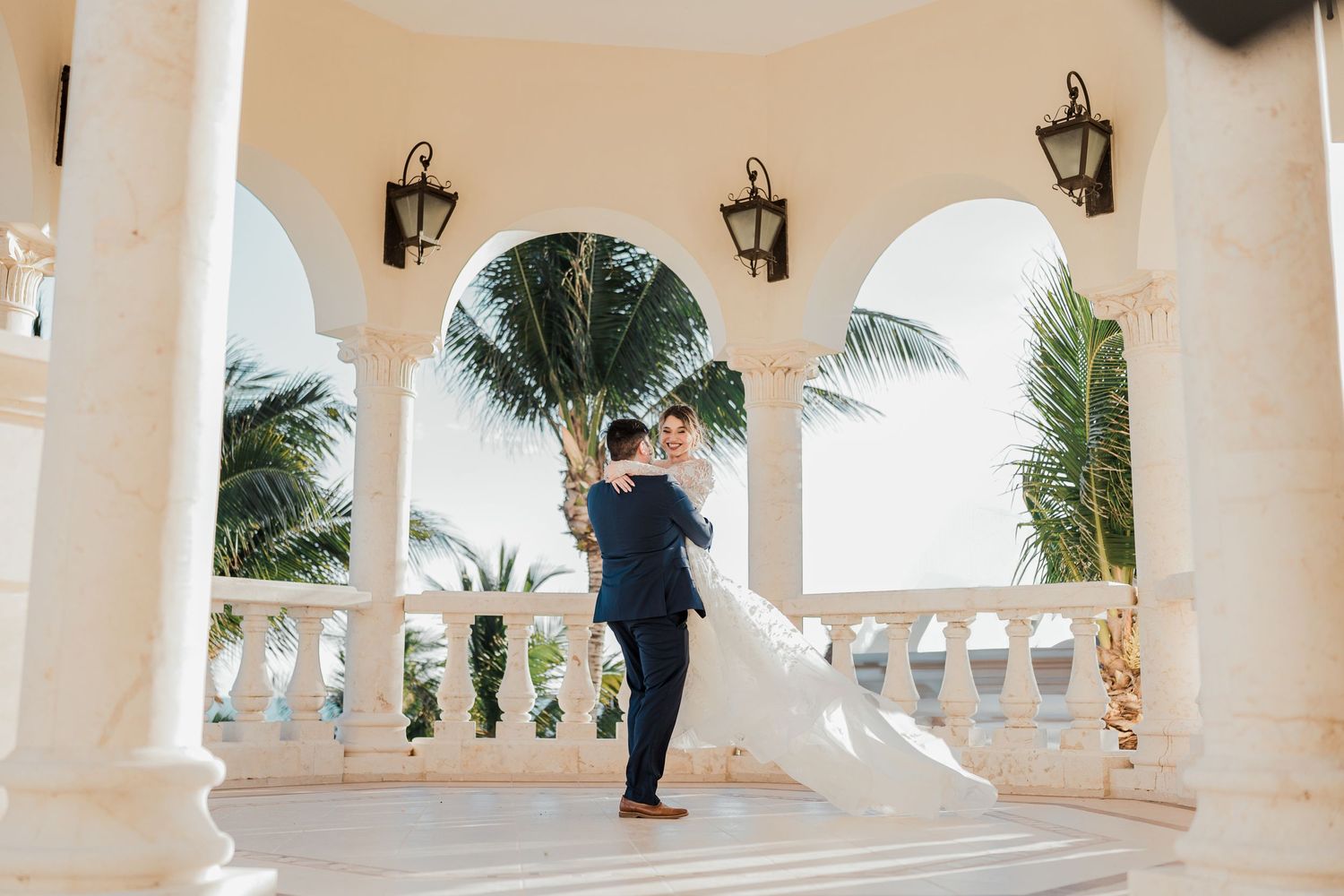 Groom lifting bride in flowing white gown under arched colonnade surrounded by tropical palm trees.
