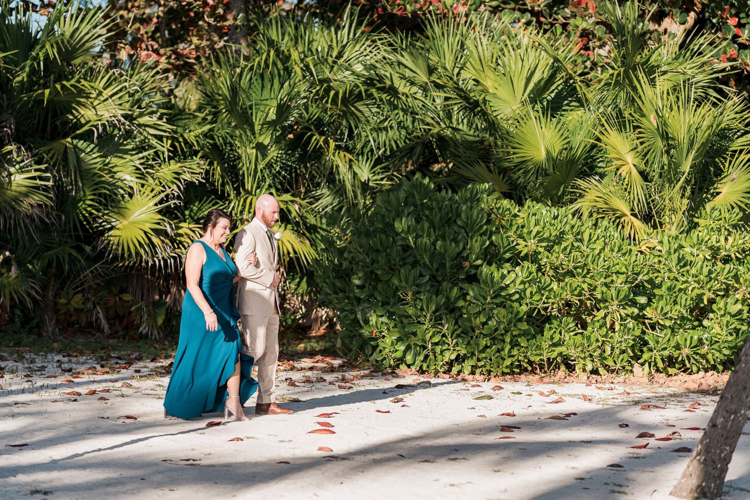 Couple in formal attire walking on sandy path surrounded by lush tropical palm trees and greenery.
