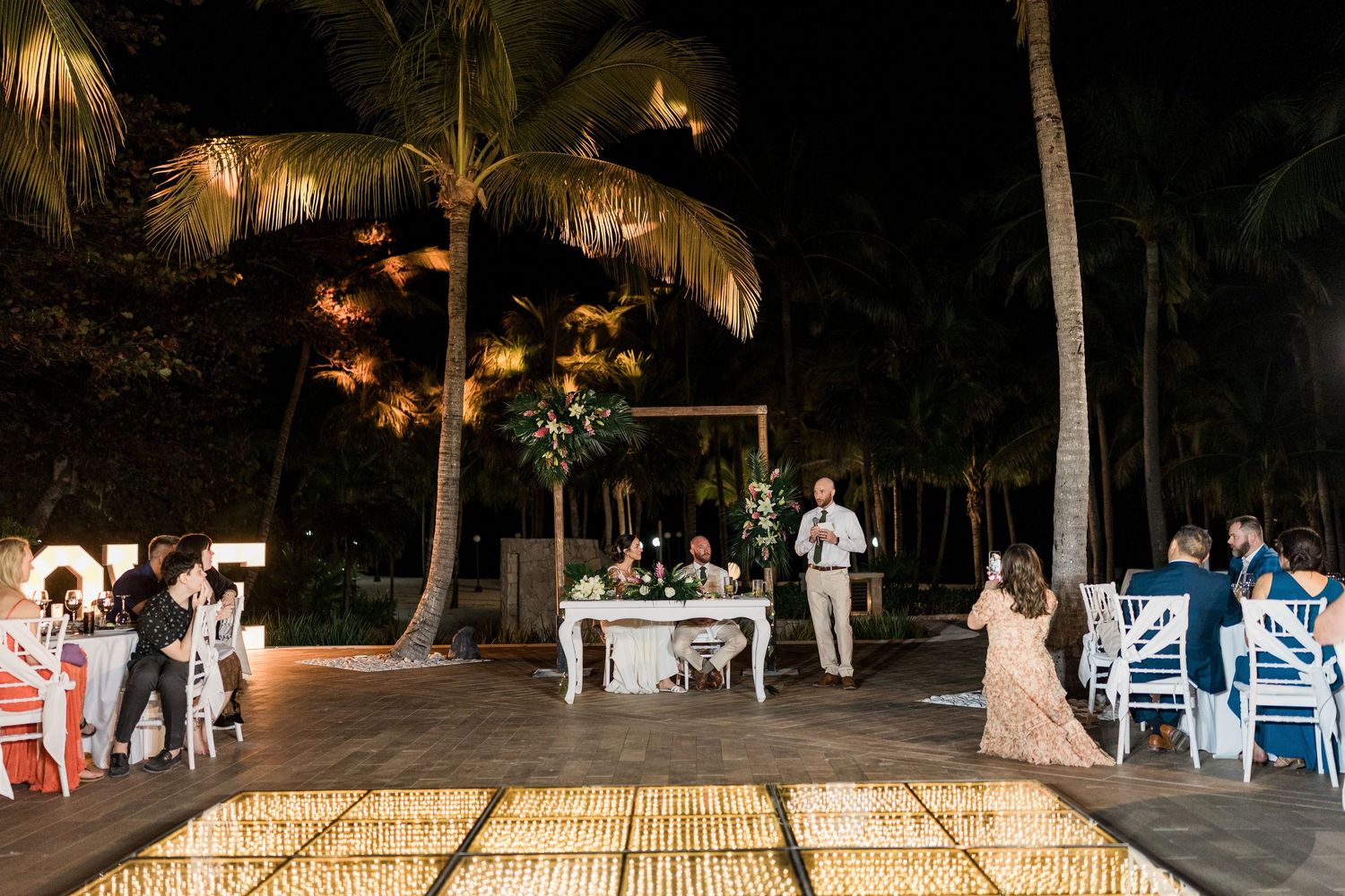 Elegant outdoor wedding reception at night with candles, palm trees, and guests gathered around a floral-decorated sweetheart table.