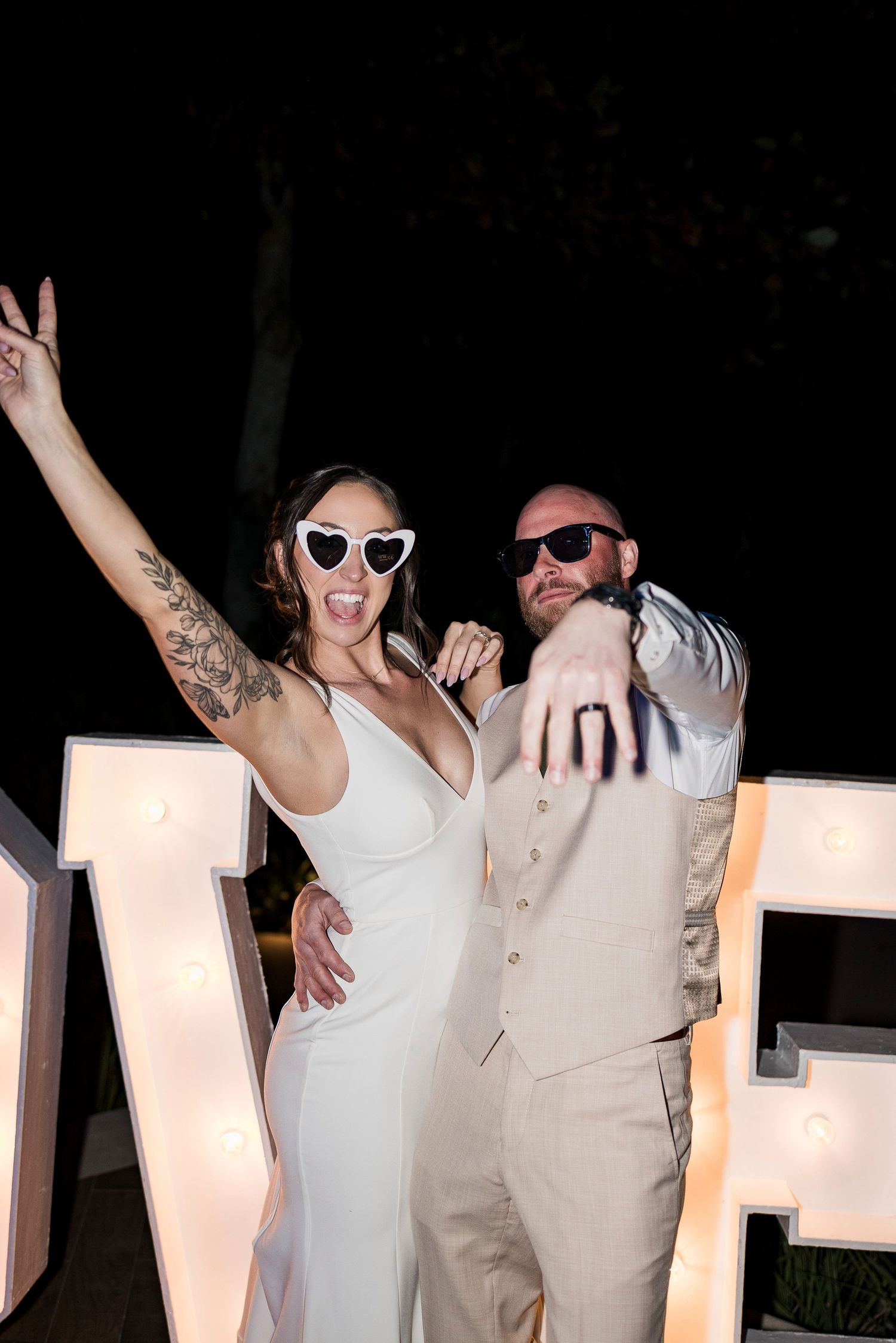 Bride and groom posing playfully in front of illuminated LOVE letters, wearing sunglasses at their outdoor evening wedding reception.
