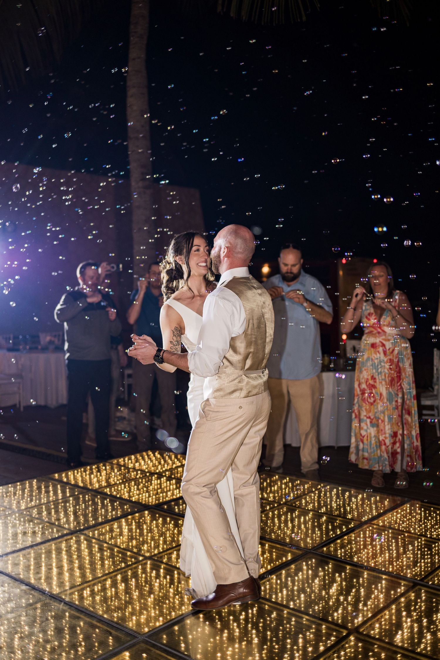 Bride and groom share first dance on glowing LED dance floor surrounded by bubbles and guests at outdoor wedding reception.