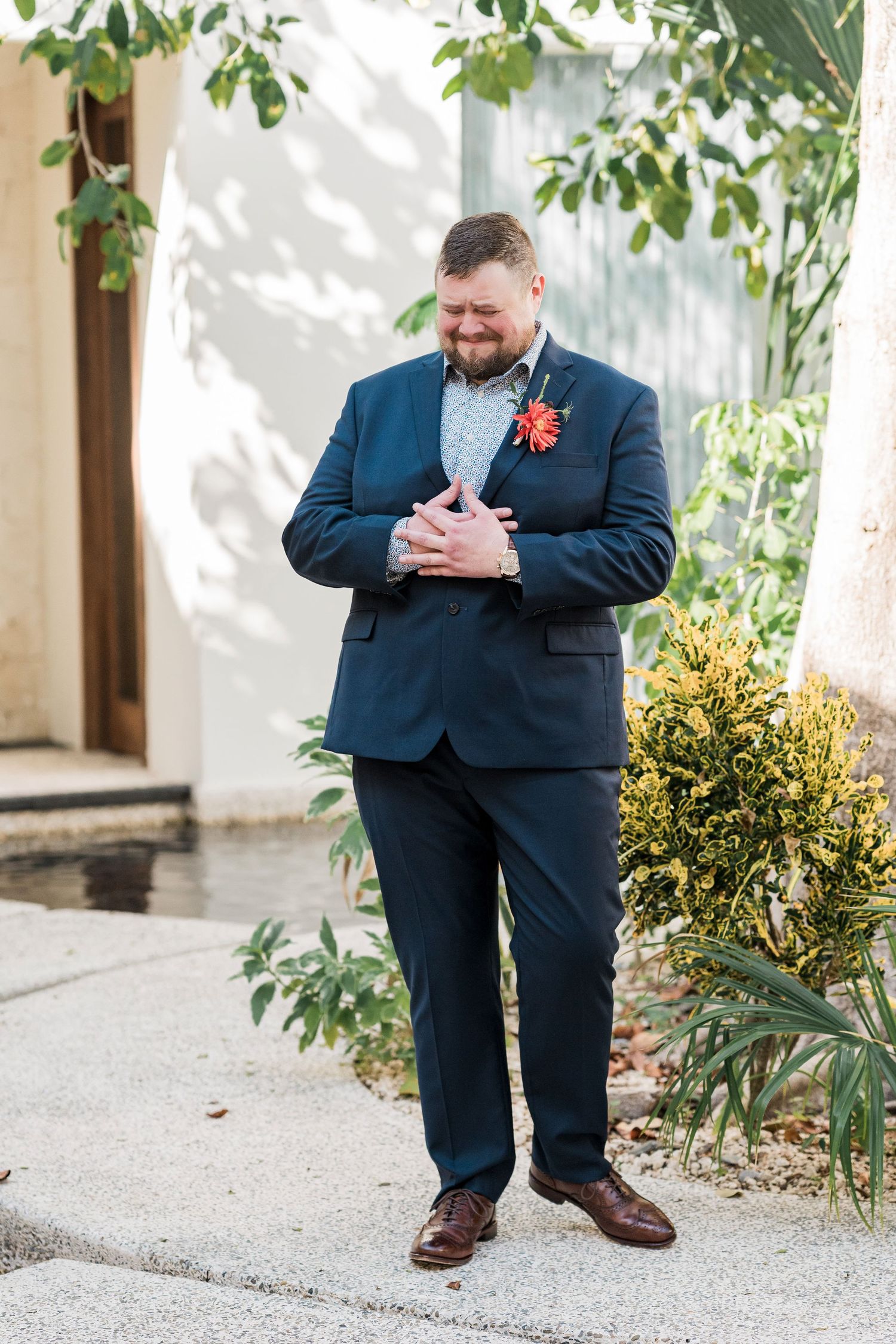 Secrets Akumal Wedding - Groom in navy blue suit with red boutonniere smiling outdoors near lush greenery on his wedding day.