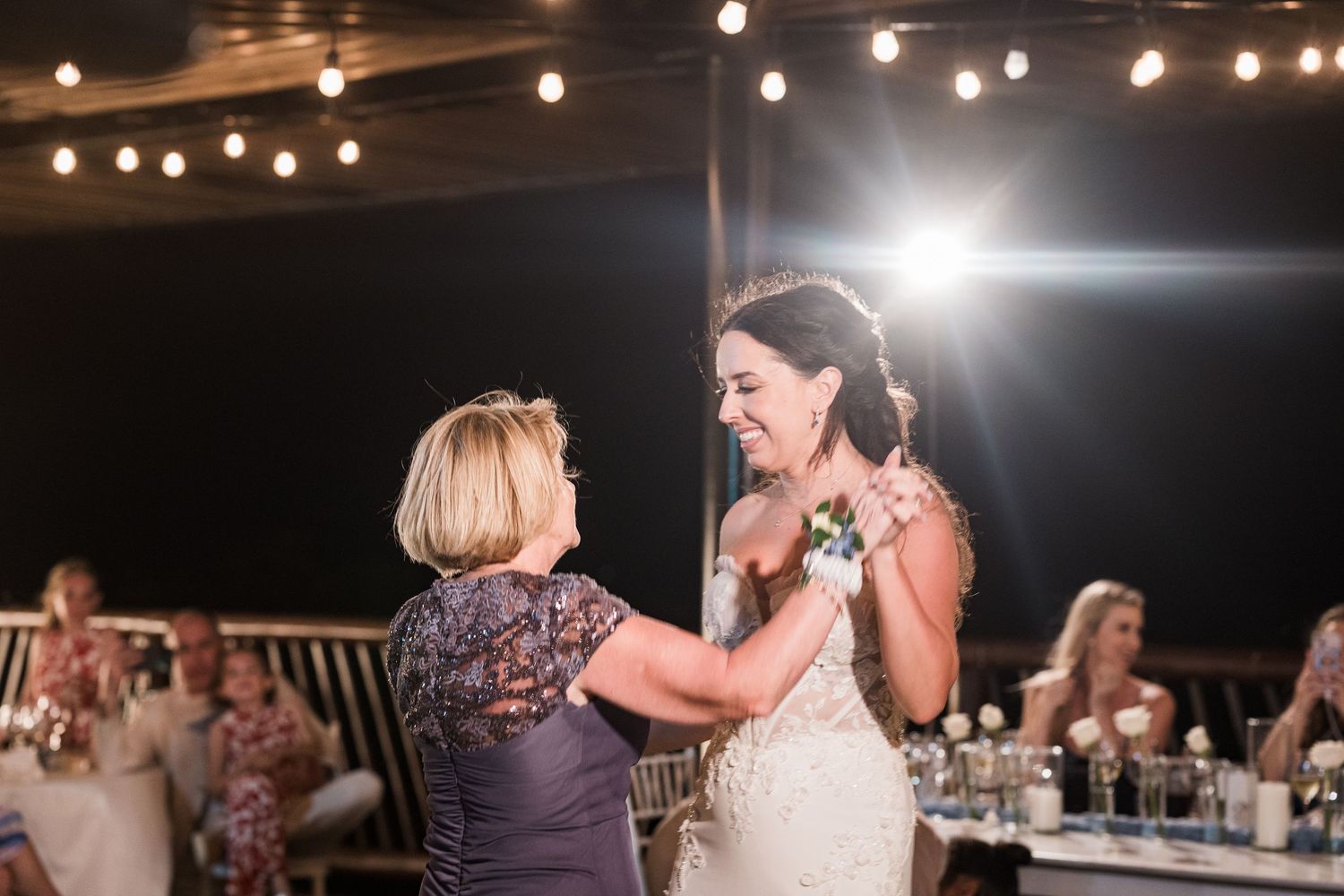 Bride and older woman share a joyful dance at an outdoor wedding reception under warm string lights.