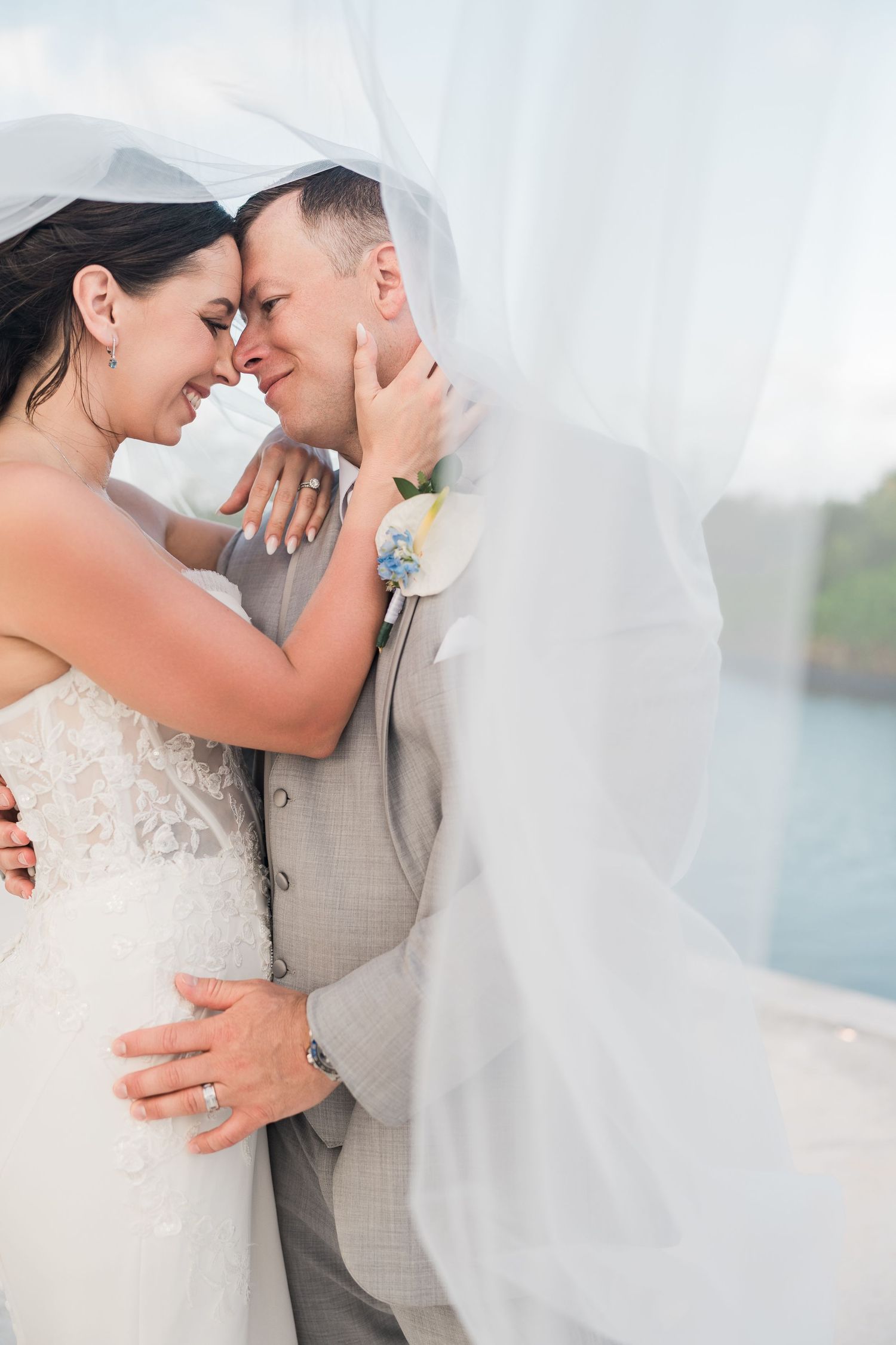 Bride and groom share a romantic moment under a flowing white veil at their outdoor waterfront wedding.