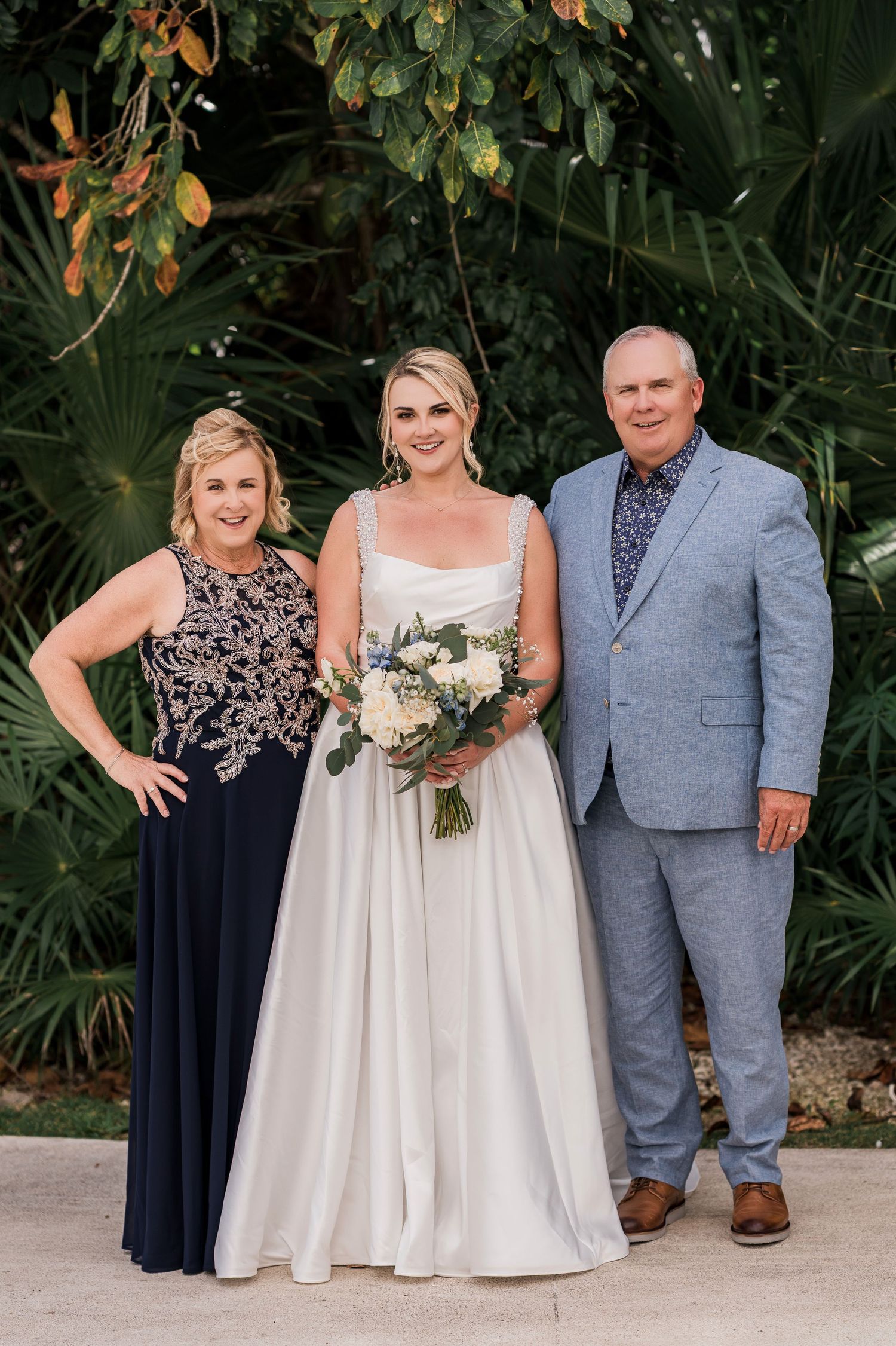 Bride in white gown holding bouquet poses with parents in tropical garden setting at outdoor wedding.