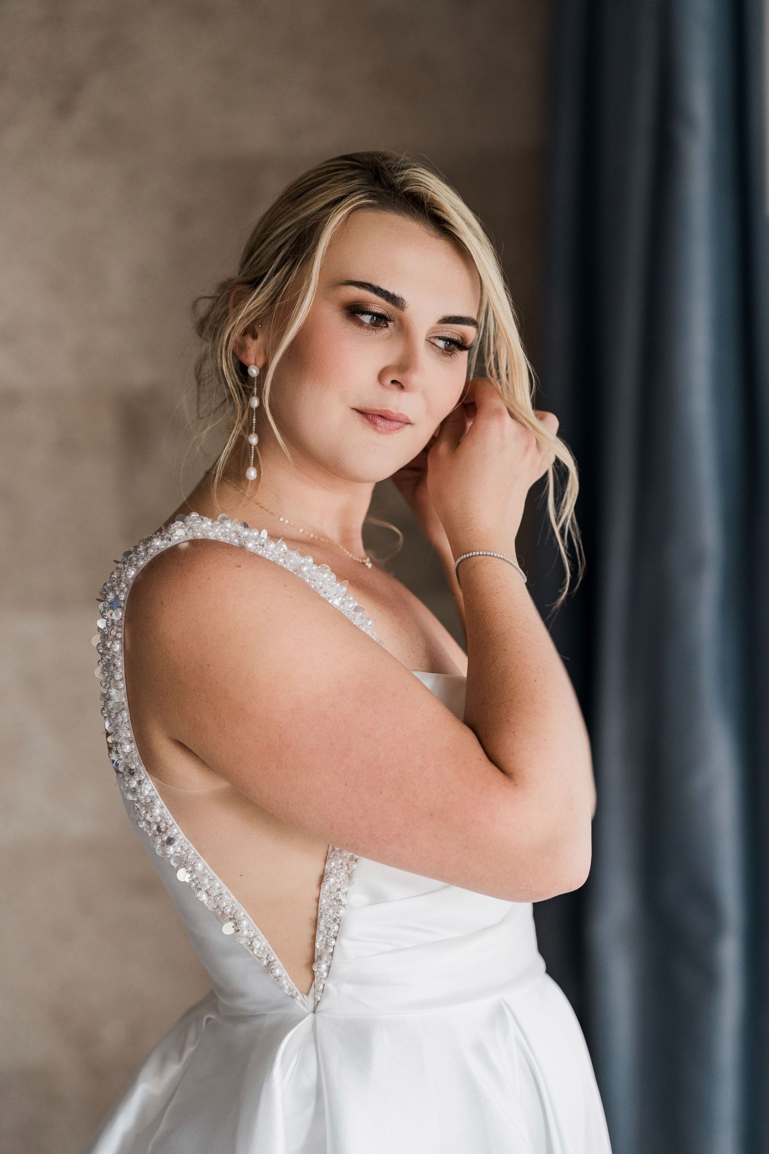 Elegant bride in a white beaded V-neck wedding gown with crystal straps, wearing drop earrings, posing gracefully indoors.