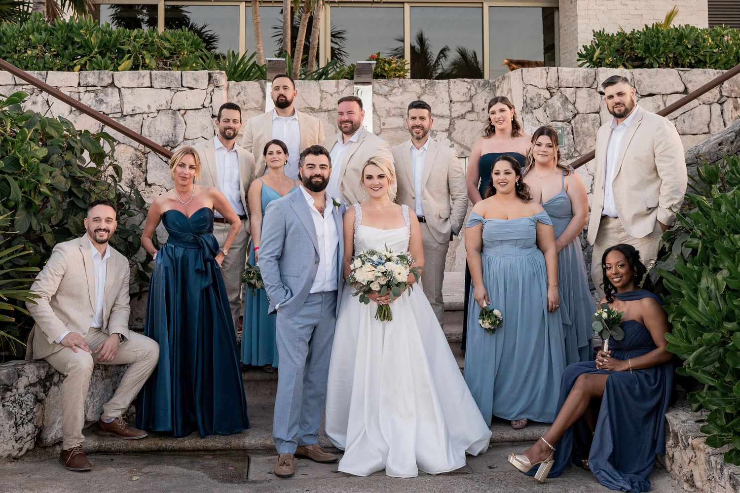 Wedding party poses outdoors in blue dresses and tan suits with bride and groom at a tropical venue. Hard Rock Riviera Maya wedding photogra