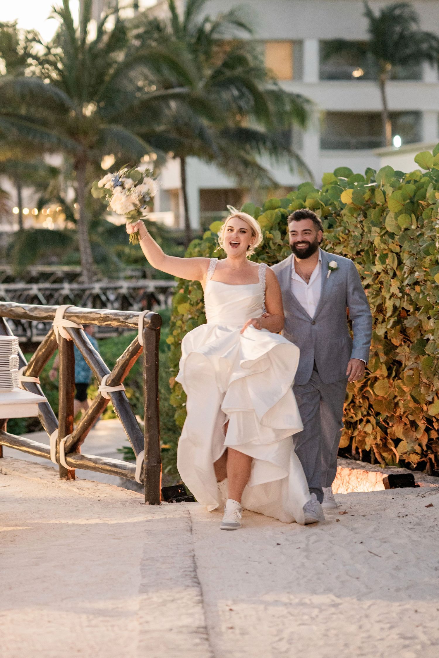 Joyful bride in white gown raises bouquet while groom in gray suit walks beside her at a tropical beach resort wedding.
