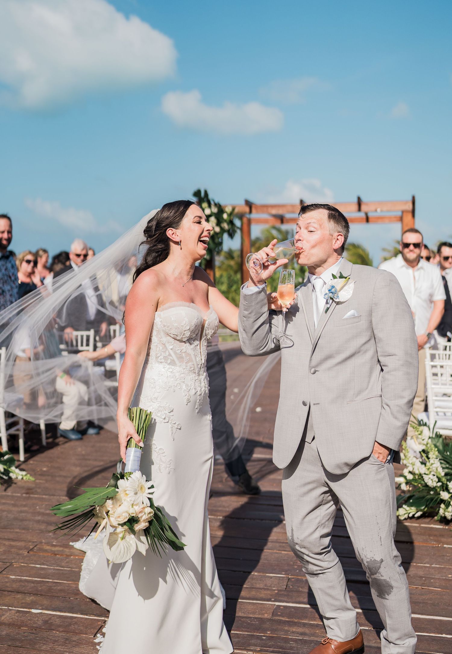Bride and groom laughing and toasting champagne after outdoor beach wedding ceremony on wooden deck.