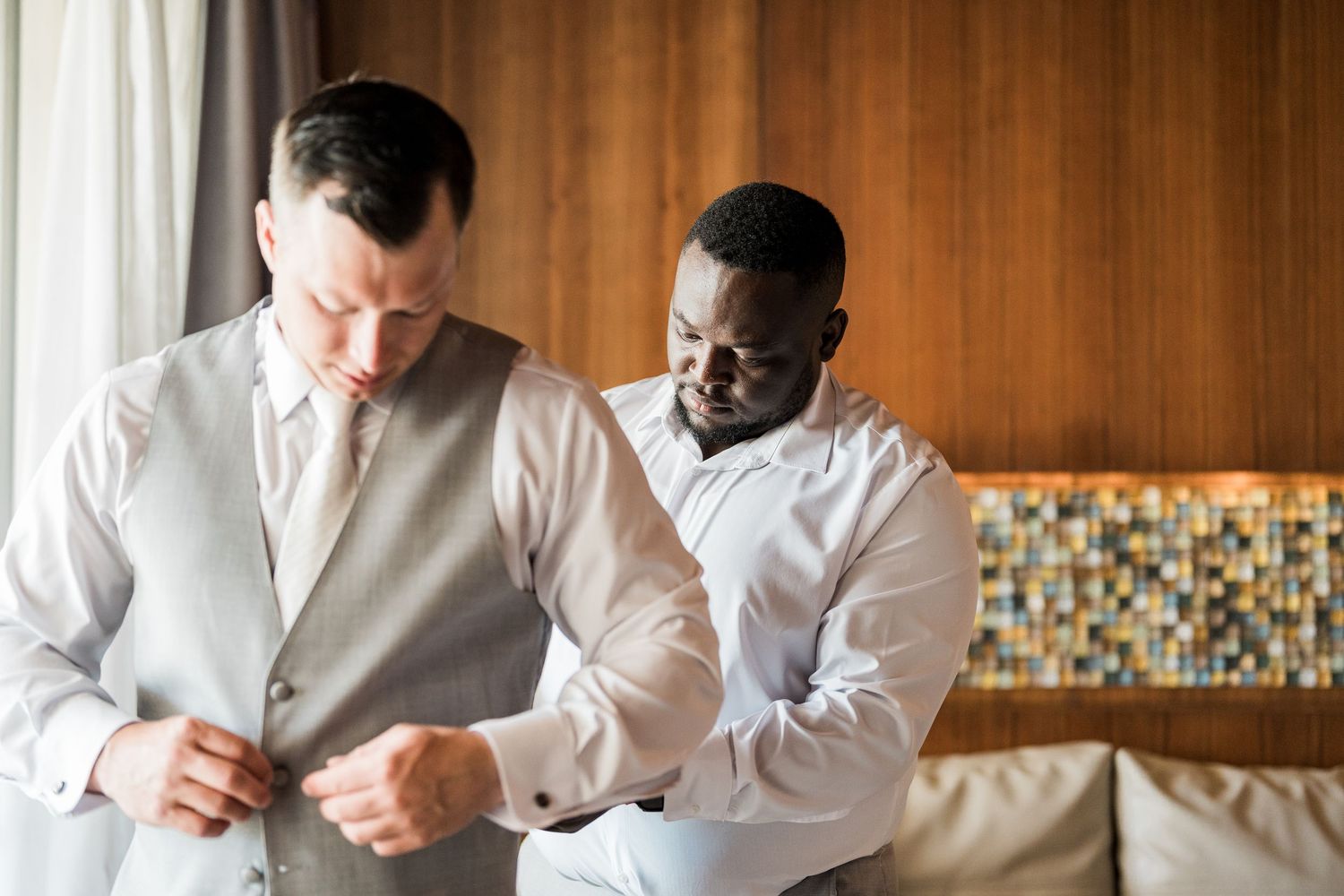Two men in formal attire help each other prepare for a wedding ceremony indoors.