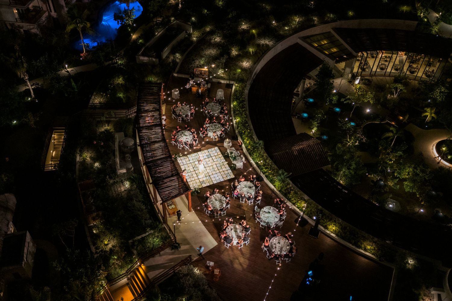 Aerial night view of an elegant outdoor wedding reception with illuminated round tables surrounded by lush gardens.