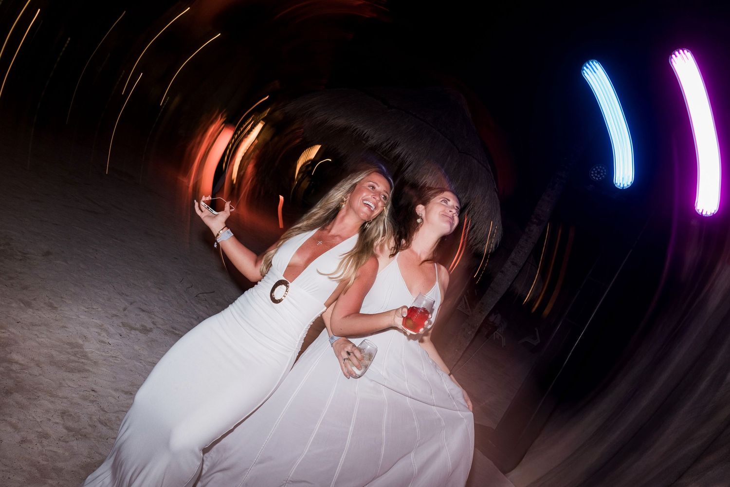 Two women in white dresses dancing joyfully at a night party with colorful light trails swirling around them.