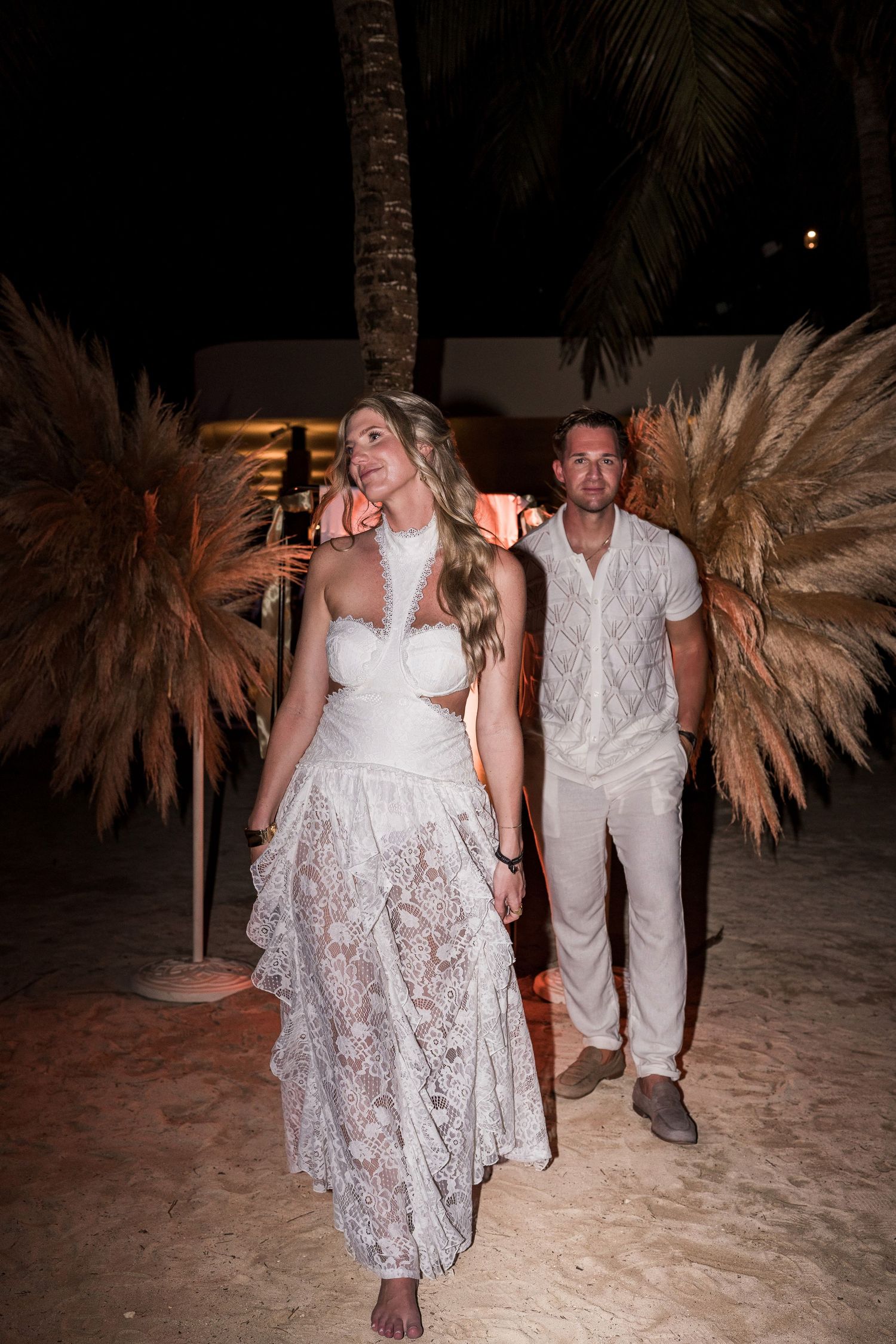 Couple in white outfits walking on sandy beach at night with pampas grass decor and palm trees.