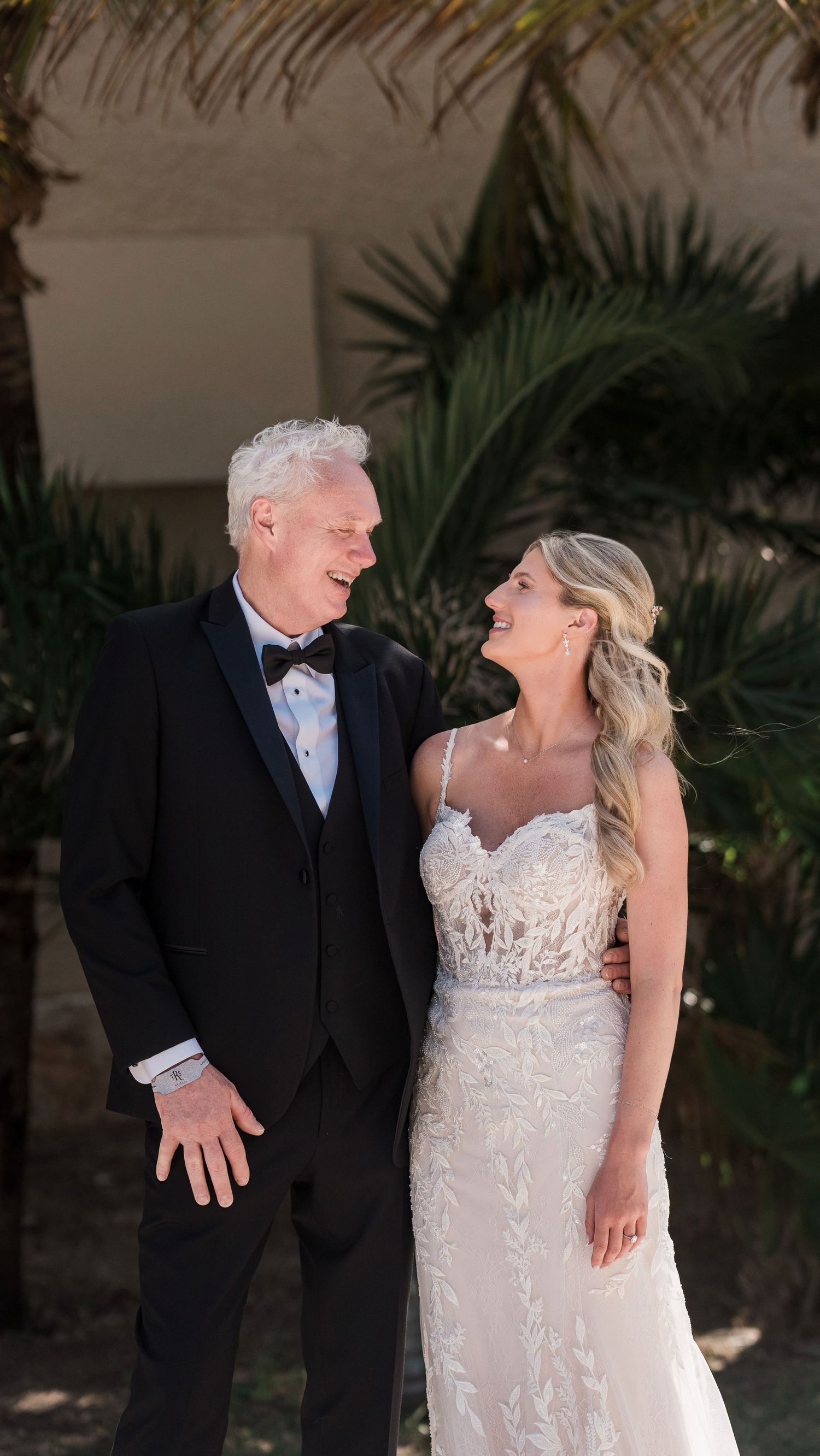 A joyful bride in lace gown and groom in black tuxedo share a loving glance outdoors with tropical palms behind them.