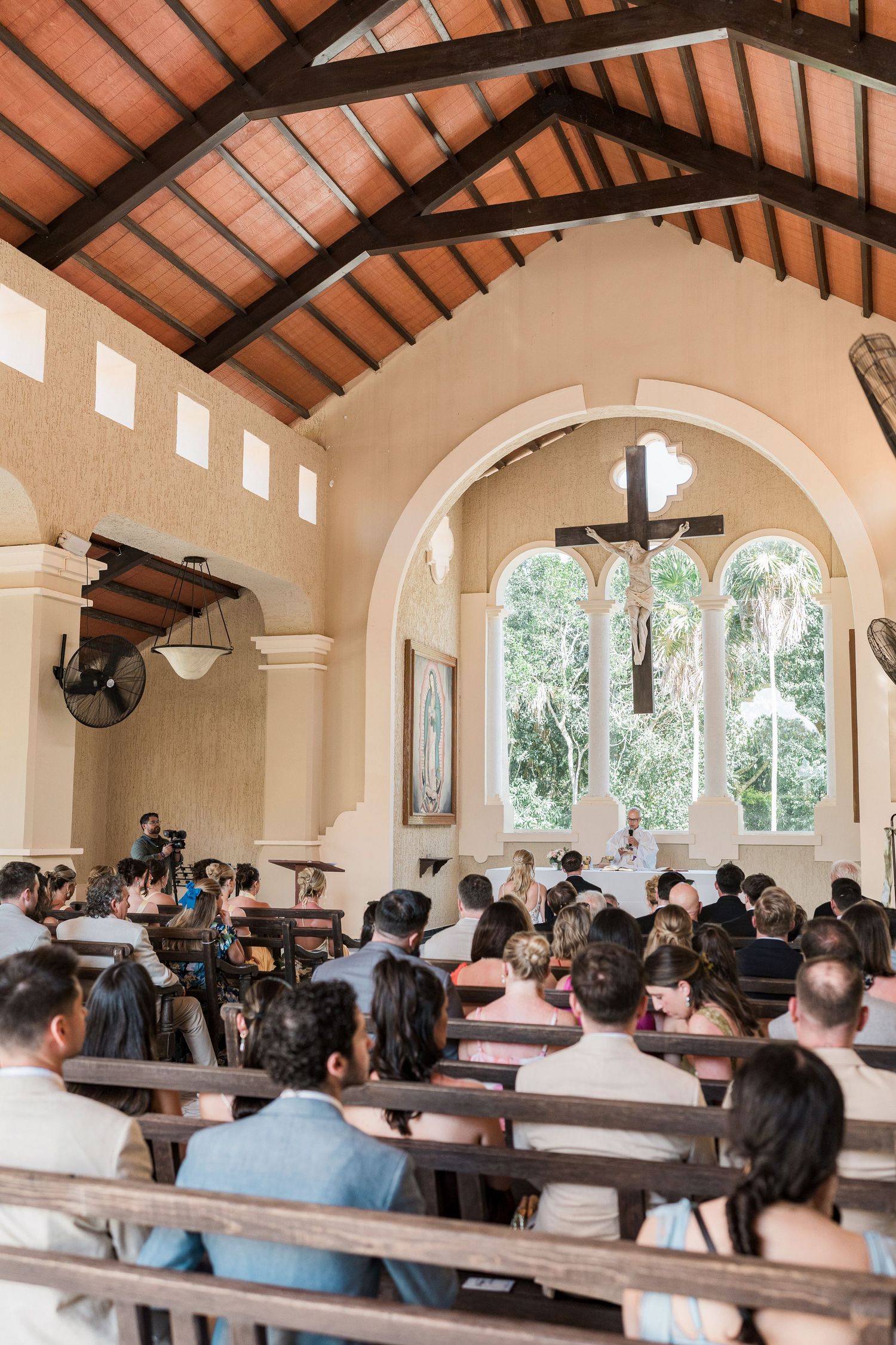 Guests seated in wooden pews inside a small chapel during a wedding ceremony, with a cross above the altar.