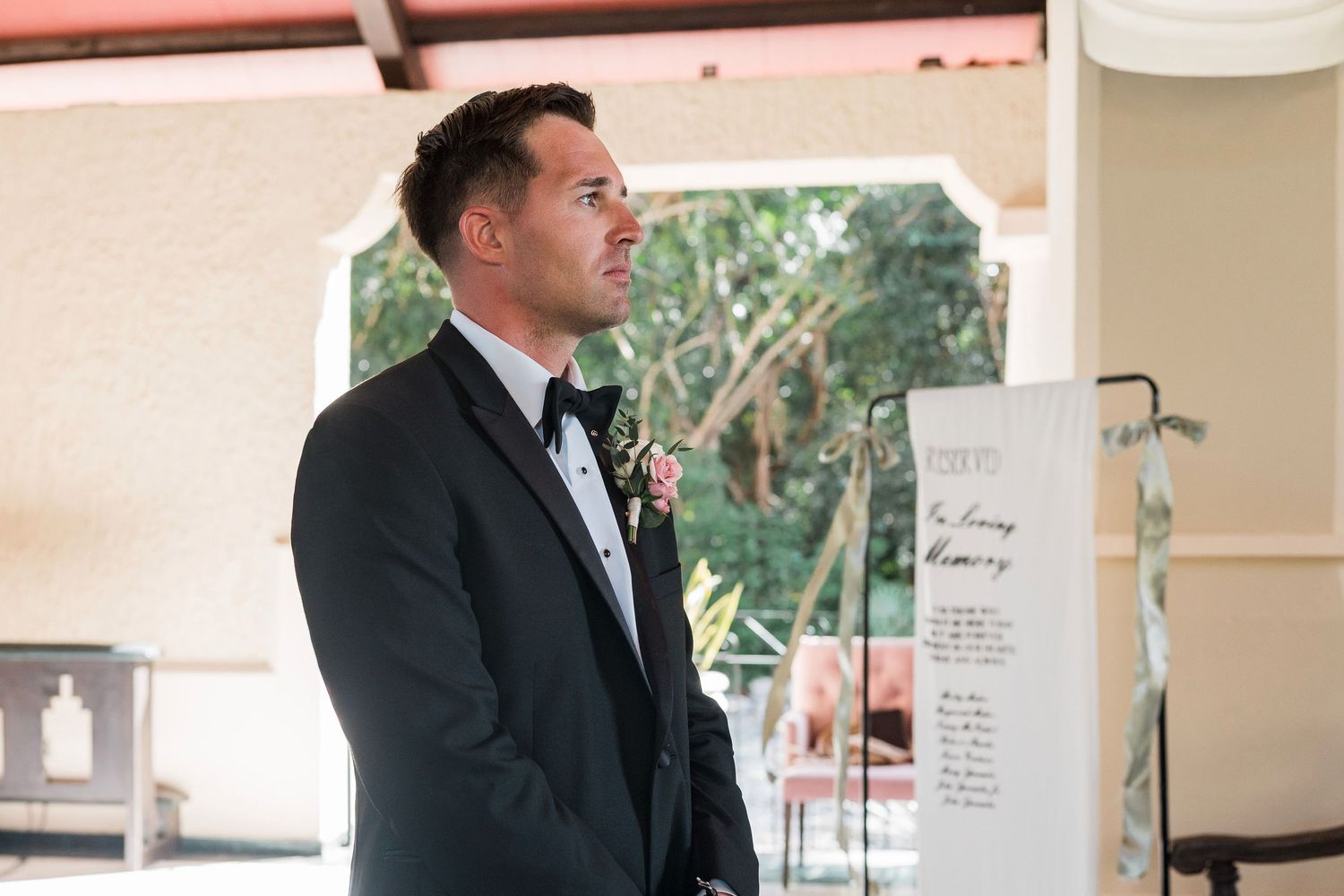 Groom in black tuxedo with boutonniere stands attentively near wedding seating chart display outdoors.