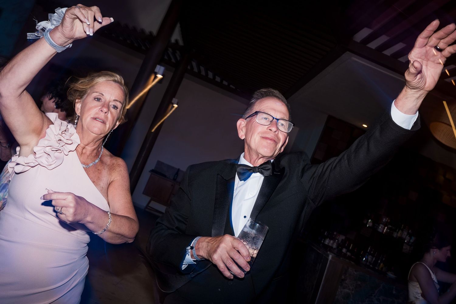 Elderly couple dancing joyfully at a formal event, woman in pink dress and man in tuxedo with bow tie.